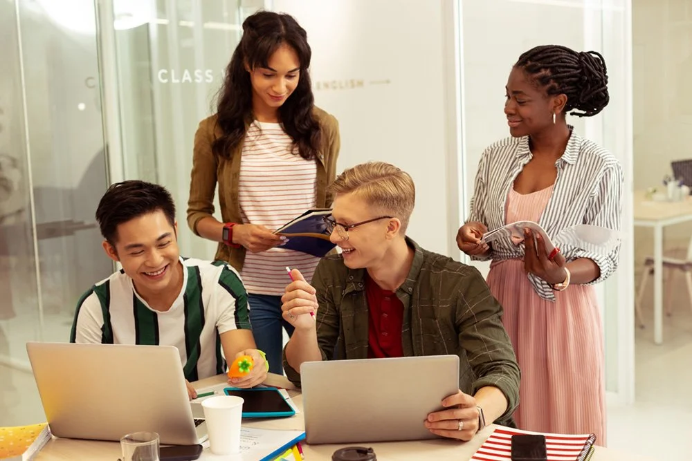 A diverse group of four young adults working together in an office or classroom, smiling and collaborating with laptops, notebooks, and tablets.