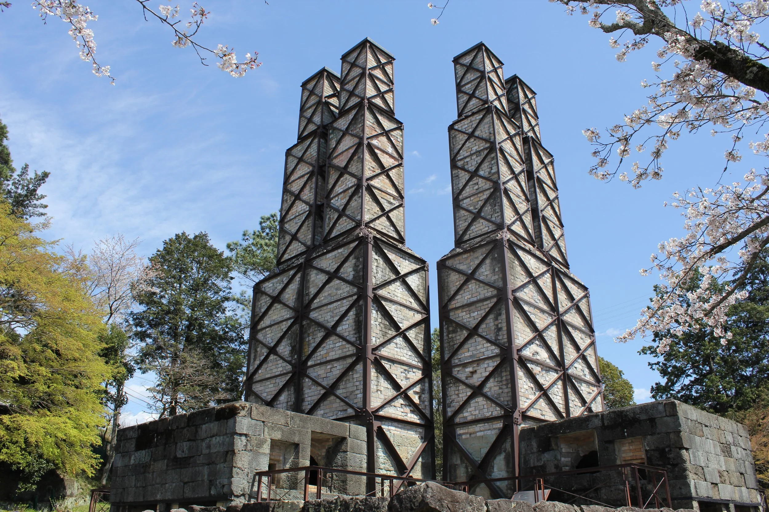 Twin brick towers of Nirayama Reverberatory Furnaces framed by cherry blossoms under a clear blue spring sky in Izu, Japan.