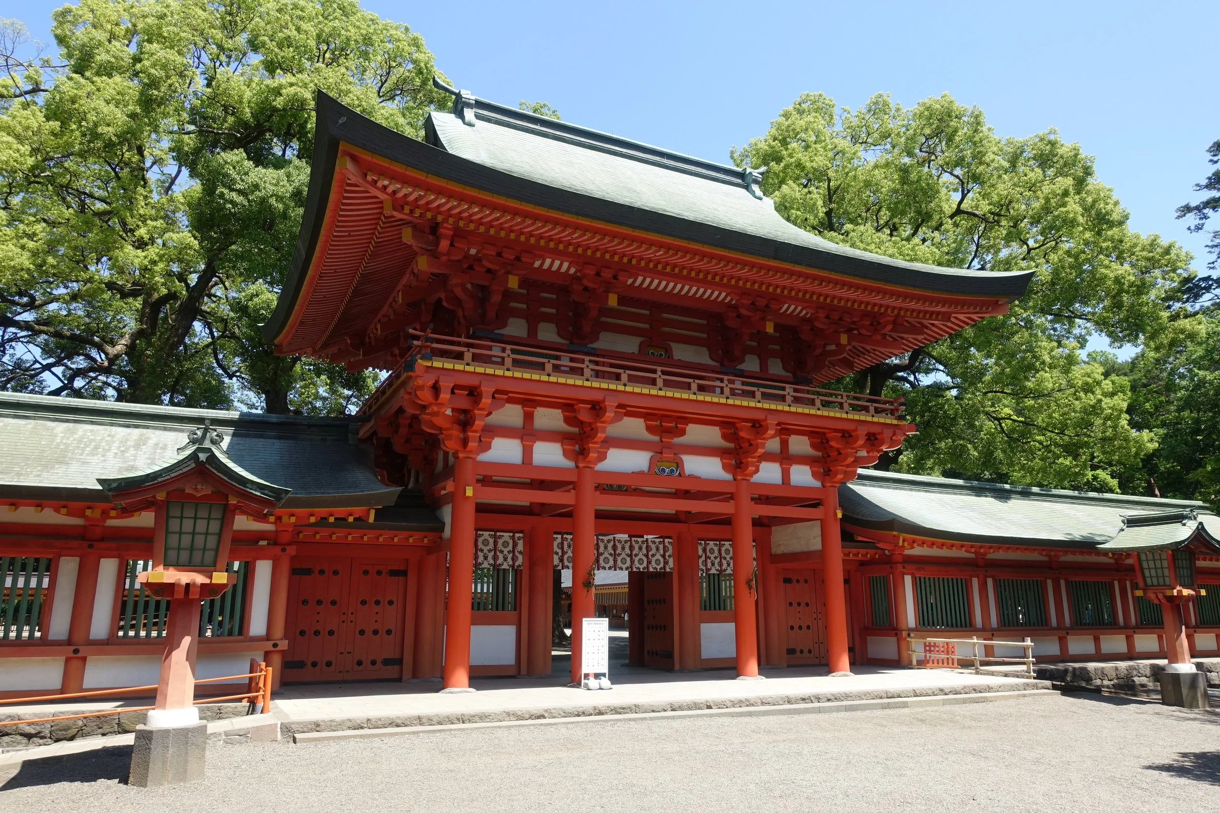 Vermilion楼門(Romon) gate of Musashi Ichinomiya Hikawa Shrine in Omiya, Saitama, framed by lush green trees under a clear blue sky.