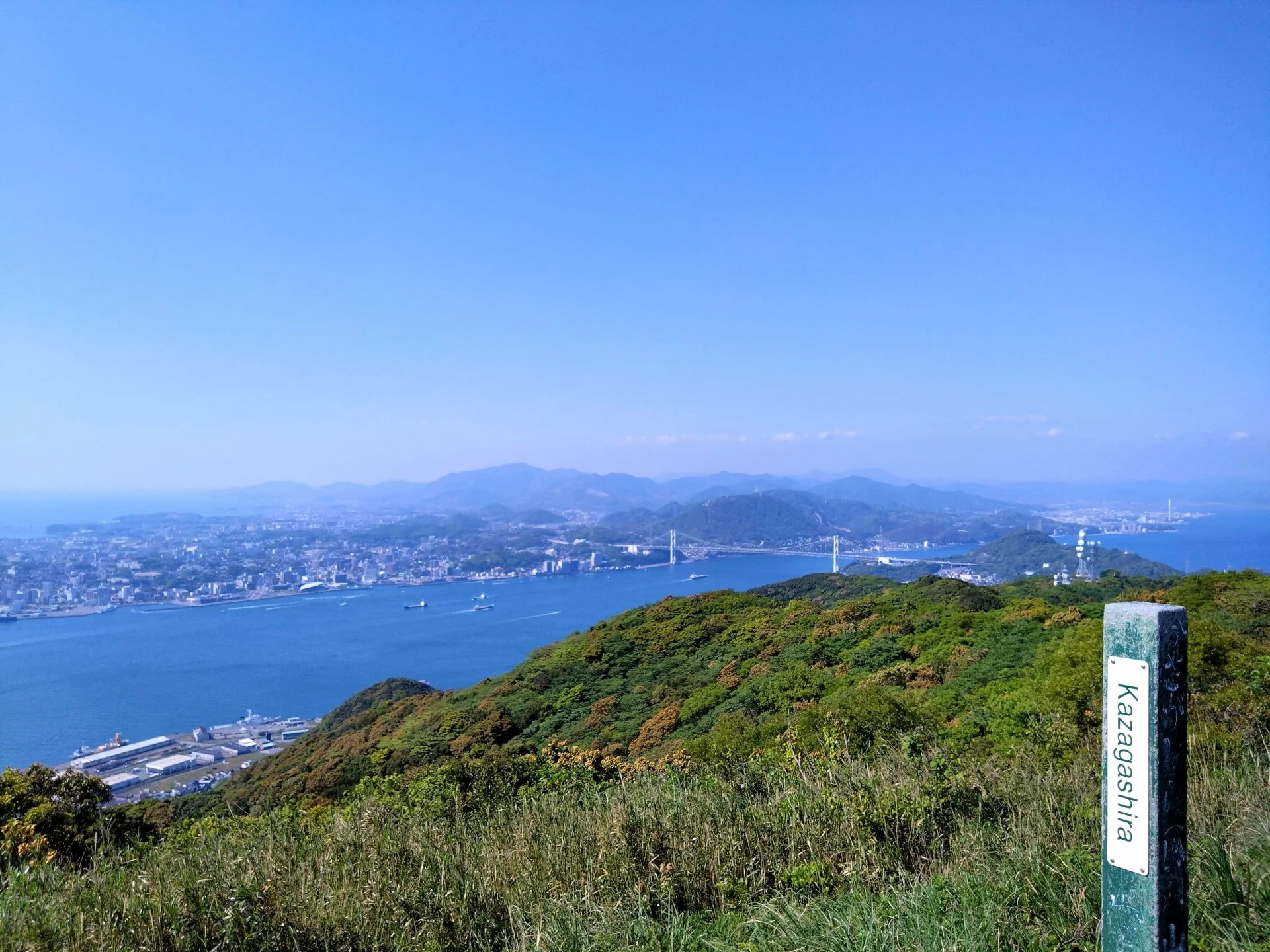 Panoramic view from Kazashi-yama in Kitakyushu overlooking the Kanmon Strait, Kanmon Bridge, and Moji coastline under a clear blue sky.