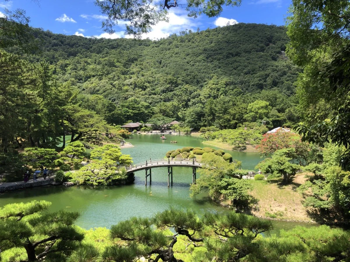 Ritsurin Garden in Takamatsu — a wooden bridge arches over the South Pond, framed by pruned pines and the forested slopes of Mt. Shiun.