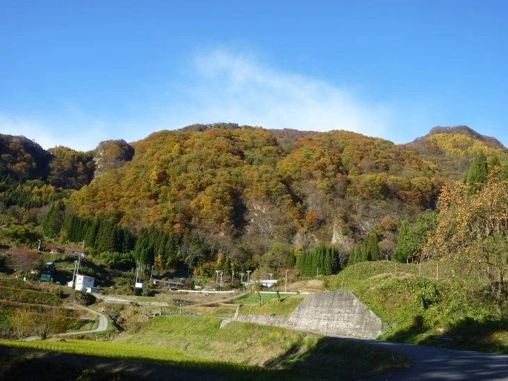 Autumn-colored slopes of Mushikura Mountain rising above a quiet satoyama village in western Nagano under clear blue skies.