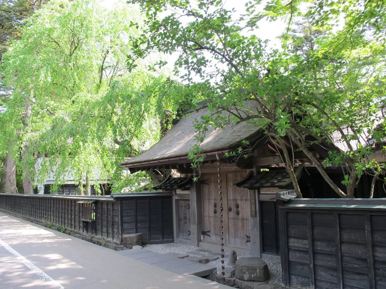 Traditional wooden yakui-mon gate at Aoyagi Samurai Manor in Kakunodate, framed by weeping willows and dappled summer sunlight.