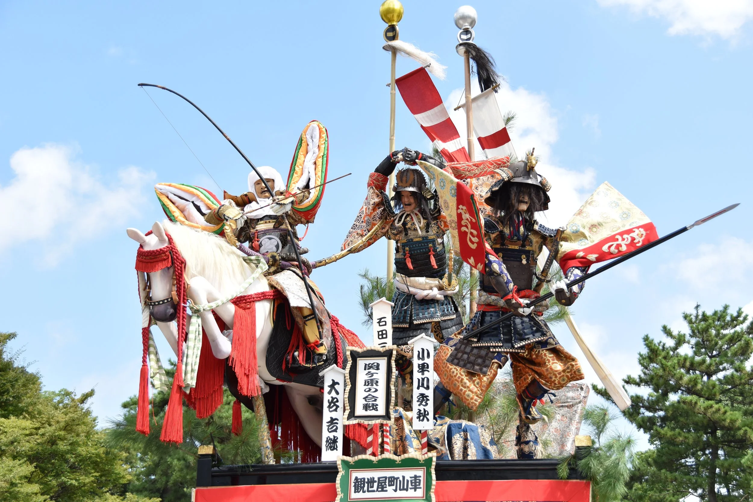 Elaborate yama festival float with life-sized samurai figures in full armor, displayed under a blue sky at the Minato Tsuruga Yama Museum in Fukui, Japan.