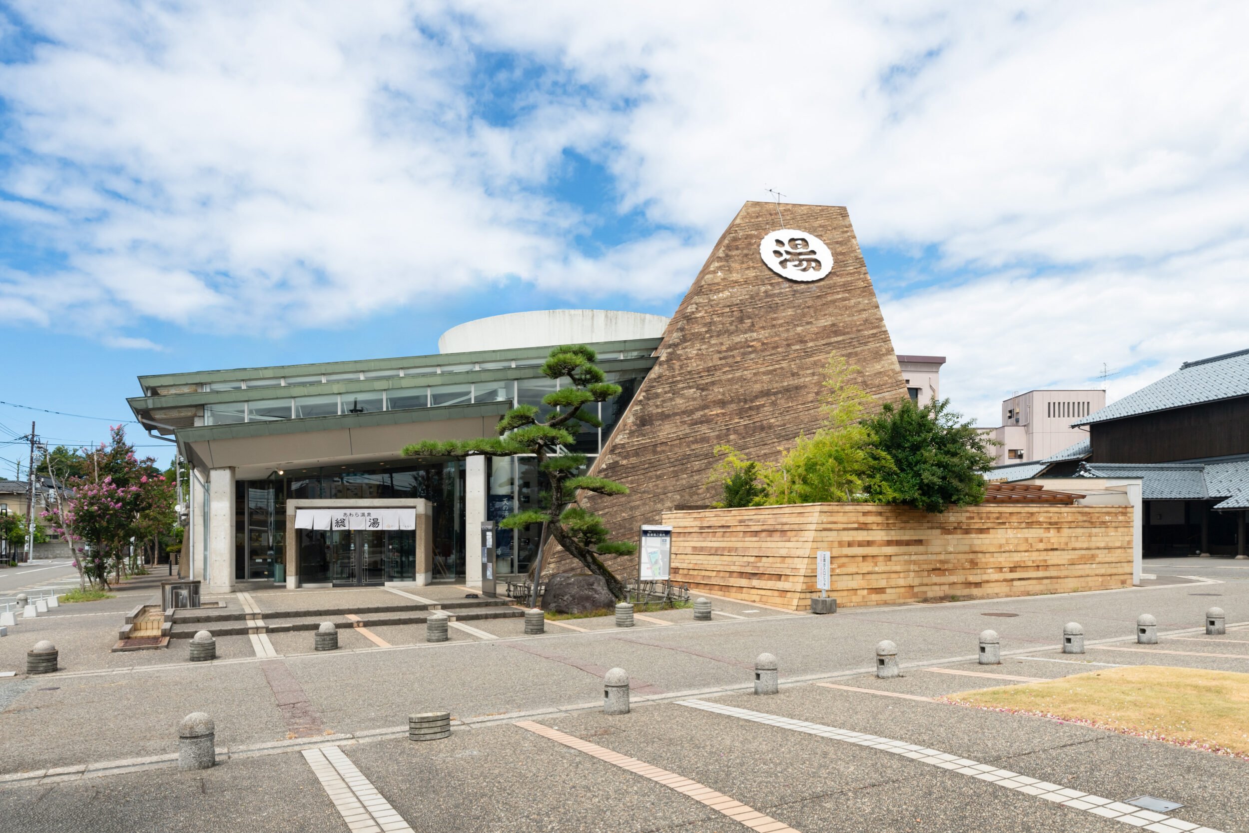 Exterior of Saintpia Awara in Fukui — a day-use onsen facility featuring a distinctive triangular monument and a spacious open plaza under a bright blue sky.