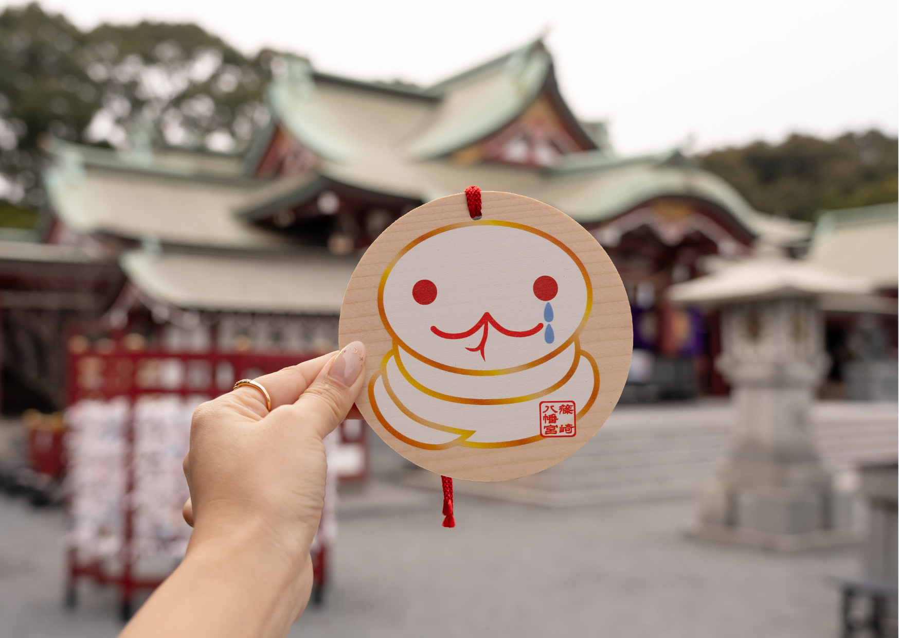Hand holding a snake-shaped ema at Shinozaki Hachimangu Shrine in Kitakyushu, with green-tiled roofs of the sacred hall softly blurred behind.