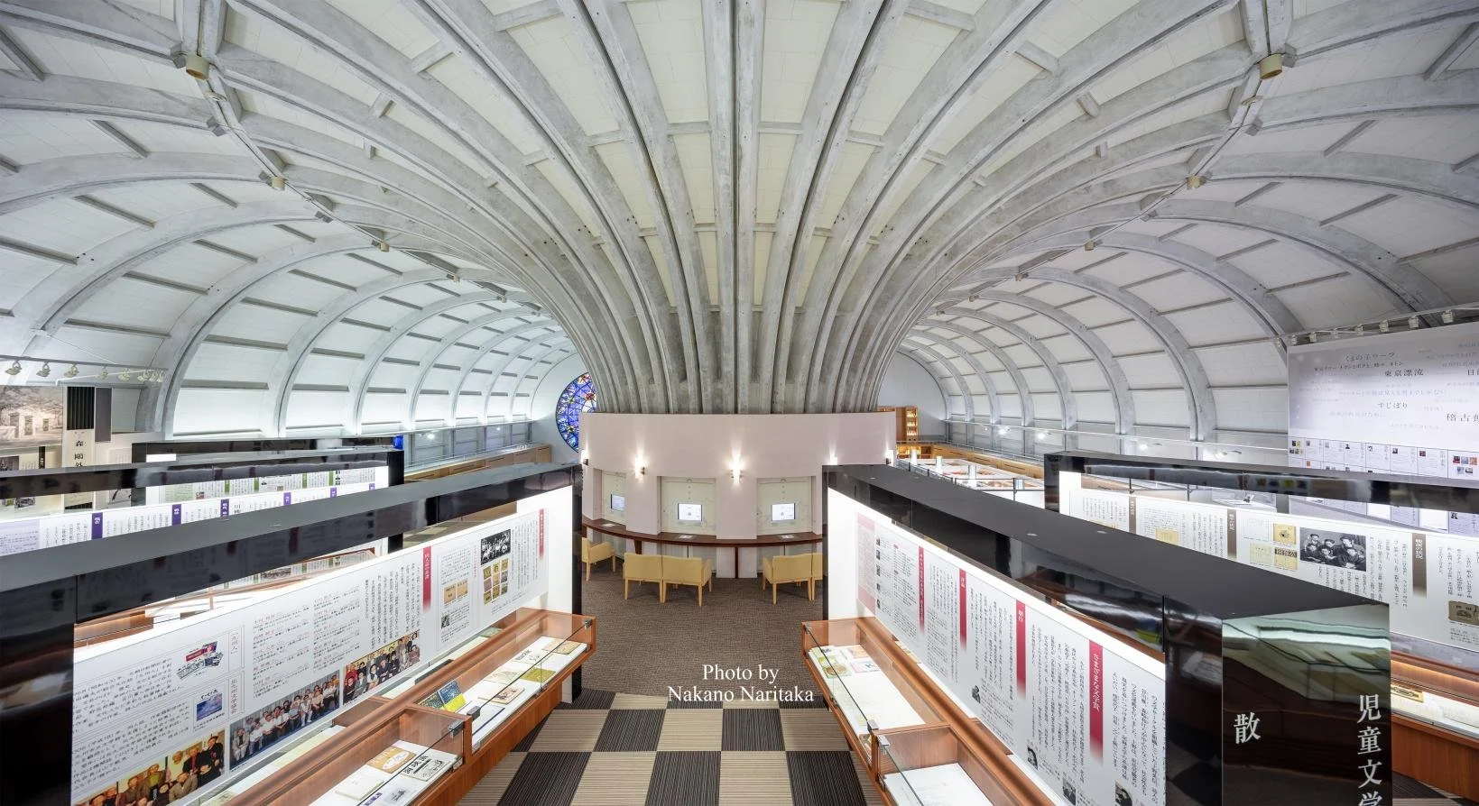 Fan-vaulted concrete ceiling and stained glass by Arata Isozaki inside the Kitakyushu Literature Museum, overlooking illuminated exhibits of Japanese literary works.
