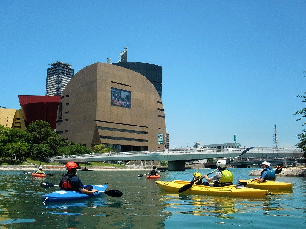 Kayakers paddling on the Murasaki River beneath a bridge with Kokura's modern skyline behind — a rare urban river experience in Kitakyushu.
