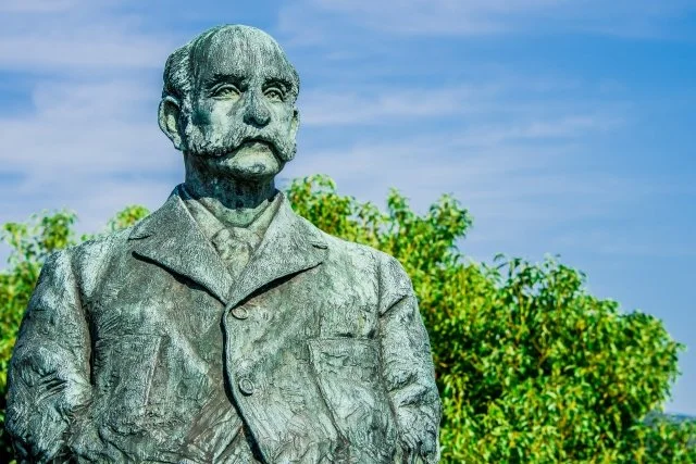 Weathered bronze statue of Scottish merchant Thomas Blake Glover in Nagasaki, set against blue sky and green trees — a symbol of Japan's modern era.
