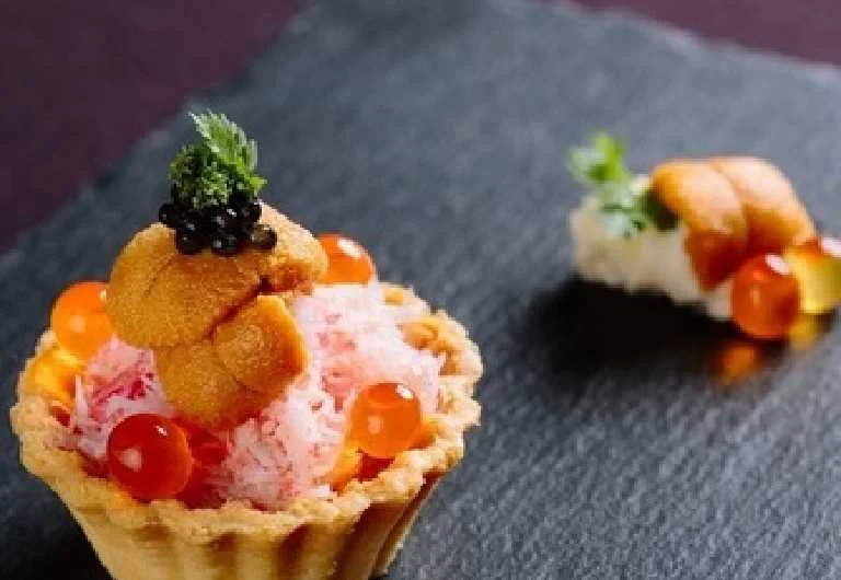 A dessert served in a tall glass with various toppings like fruit, beans, and cookies, viewed on a tray with a blurred background.