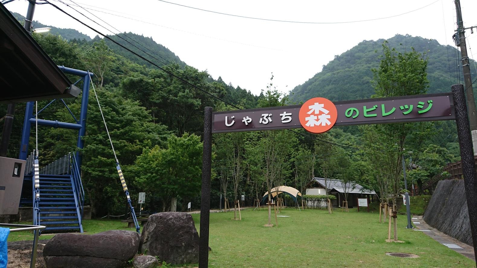 Entrance sign of Jabuchi Mori no Village campground in Miyako Town, Fukuoka, framed by green forested hills and open lawn.