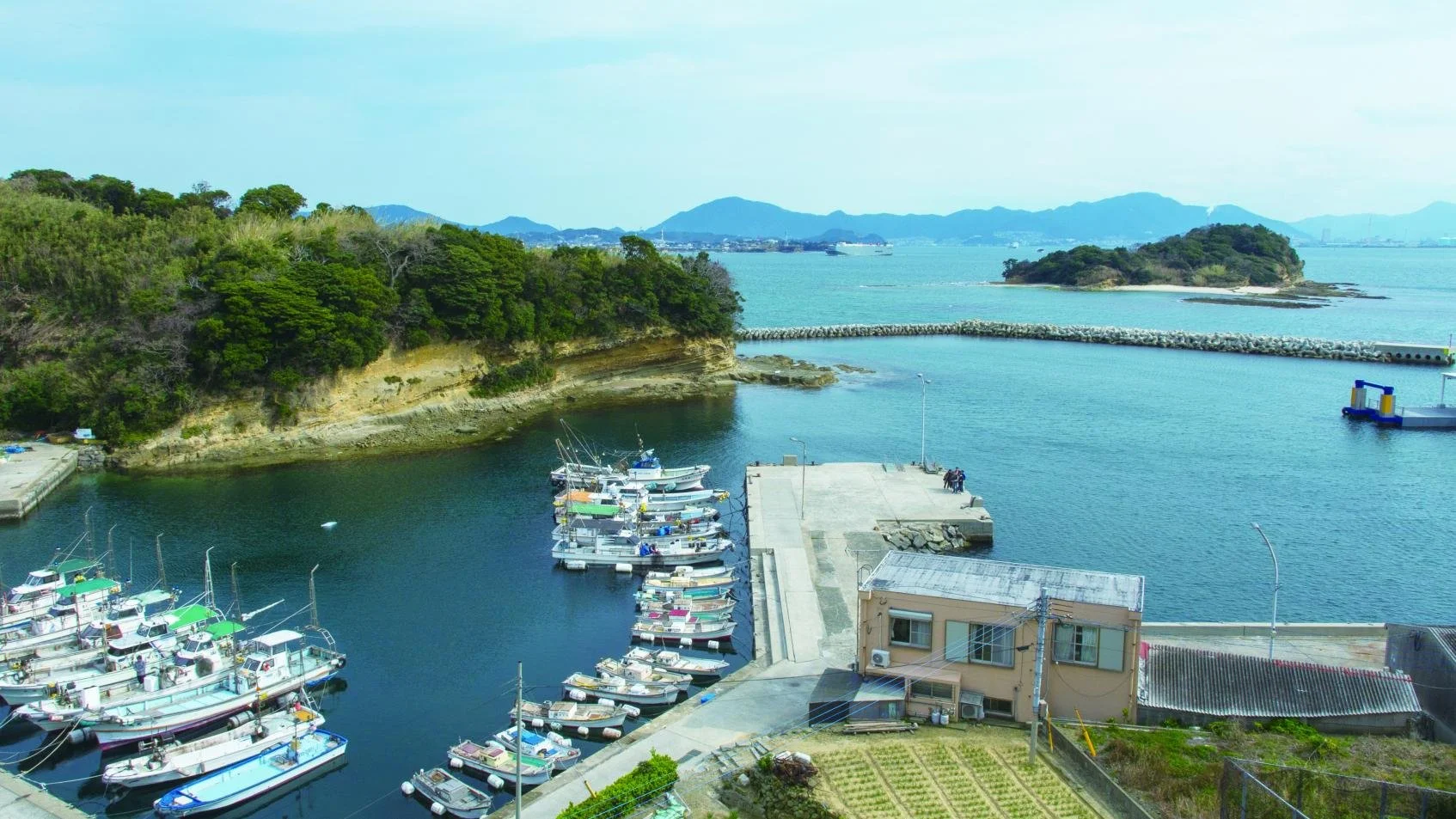 Small fishing boats moored in the quiet harbor of Umashima, a hidden island off Kitakyushu, with green hills and calm sea stretching to the horizon.