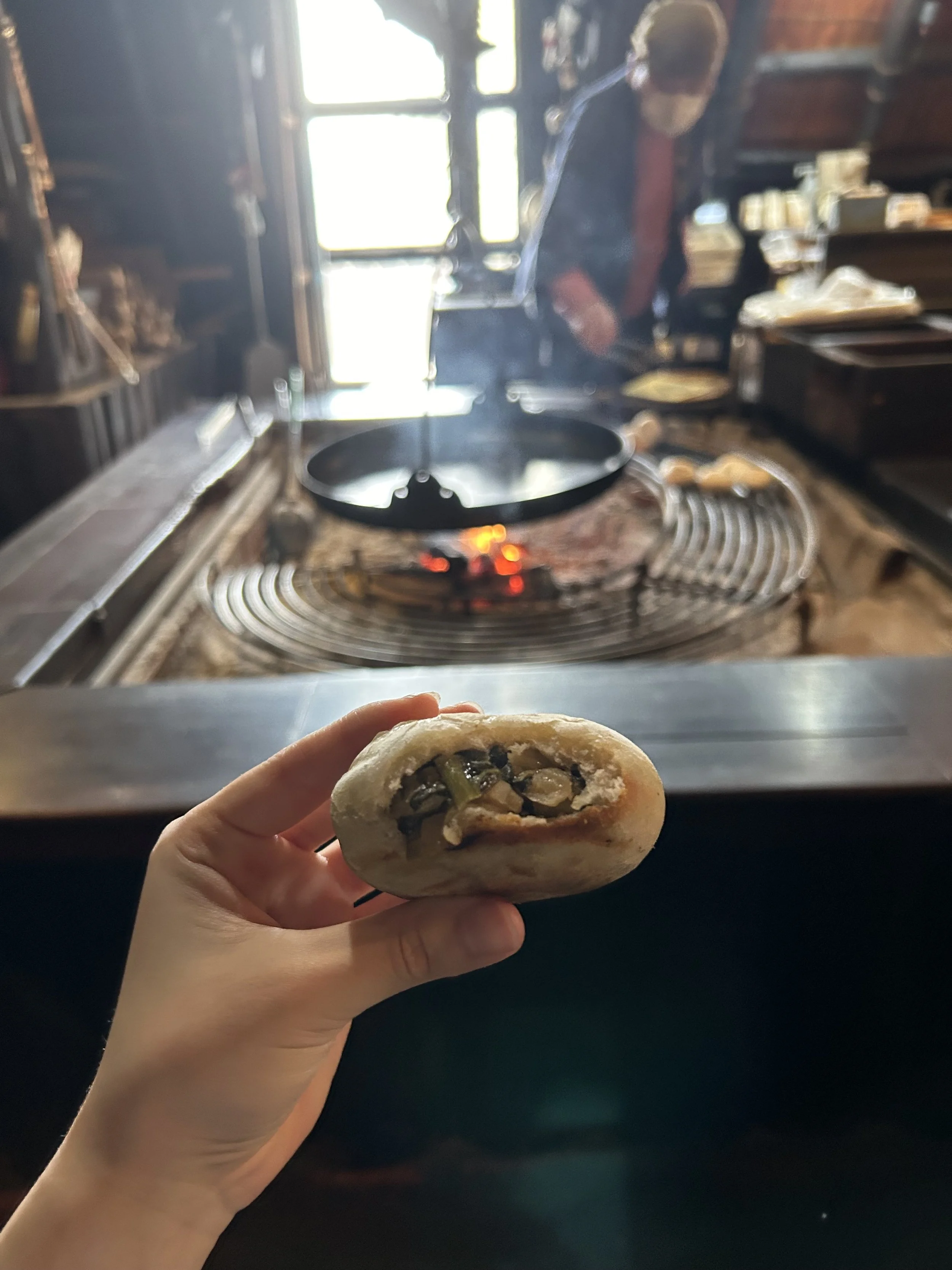 Hand holding a freshly baked oyaki bun by a glowing irori hearth at Ogawanosho Jomon Oyaki-mura in rural Nagano, Japan.