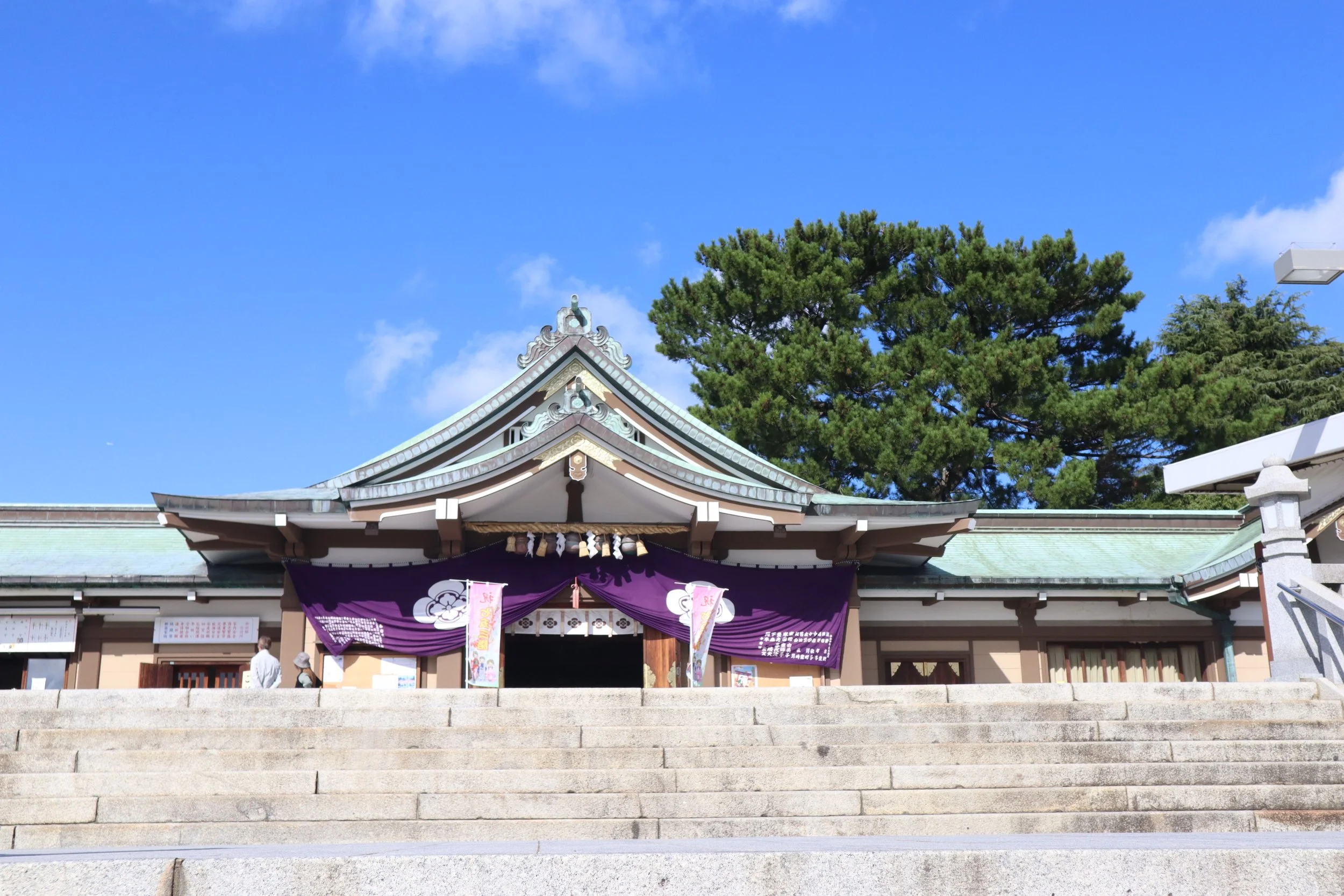 Traditional main hall of Kameyama Shrine in Shimonoseki, framed by deep purple noren curtains, a green patinated roof, and evergreen trees under a clear blue sky.