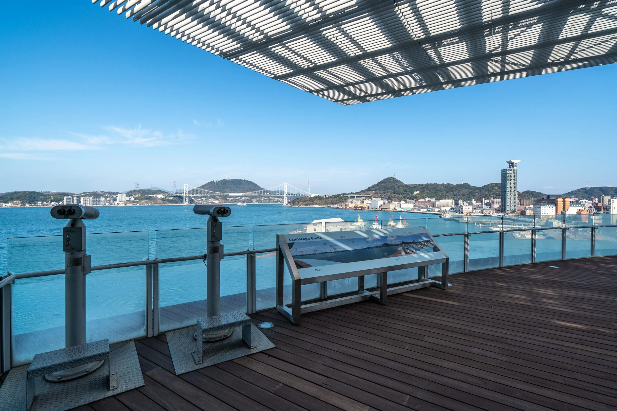 Observation deck overlooking a cityscape with water, hills, and a bridge, featuring binoculars and informational signage.