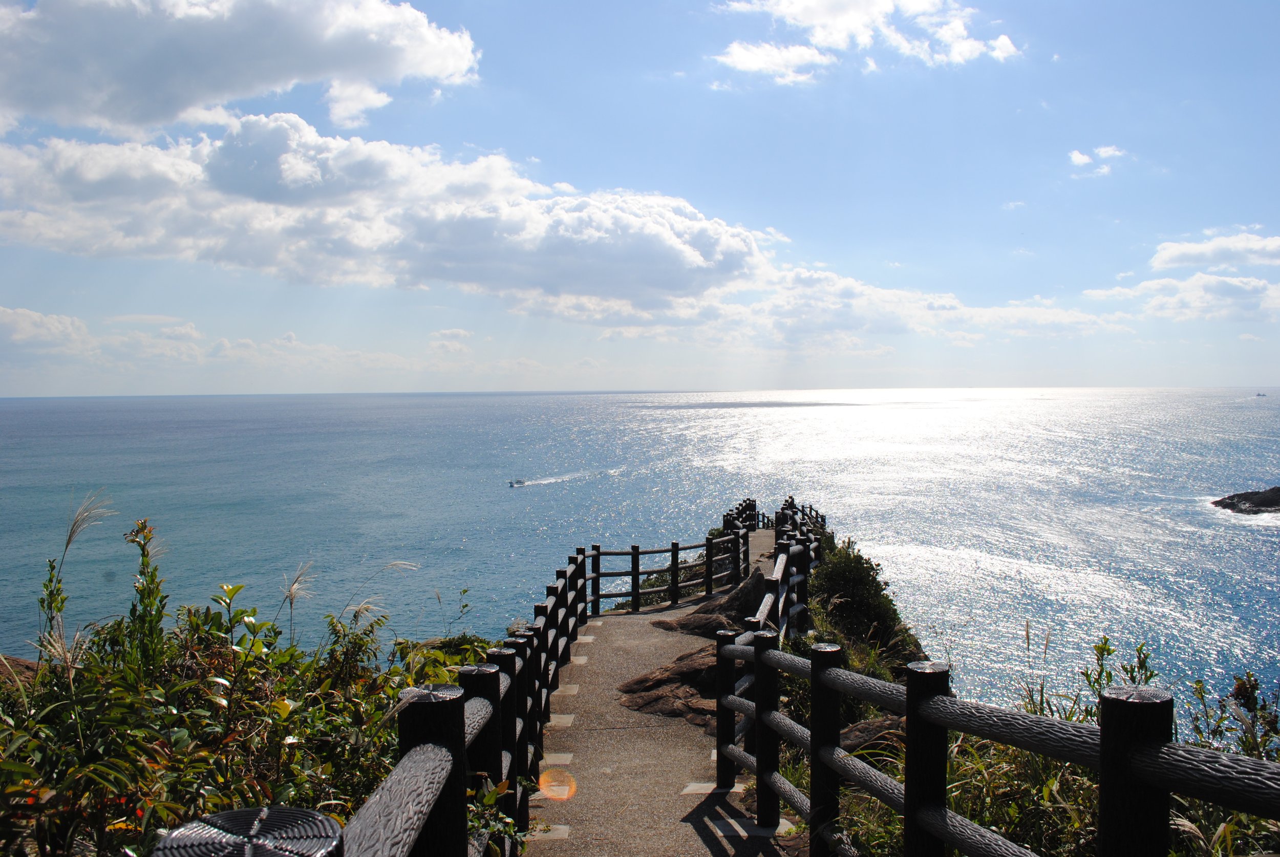 Wooden-railed clifftop path at Umagase leading toward the Pacific Ocean, with columnar basalt cliffs and glittering sea beyond — Hyuga, Miyazaki.
