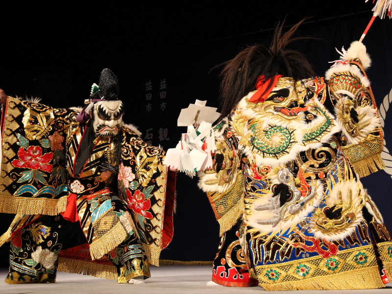Two Iwami Kagura performers in gold-embroidered costumes and ornate masks on a Masuda stage, bringing ancient Japanese myths to life.