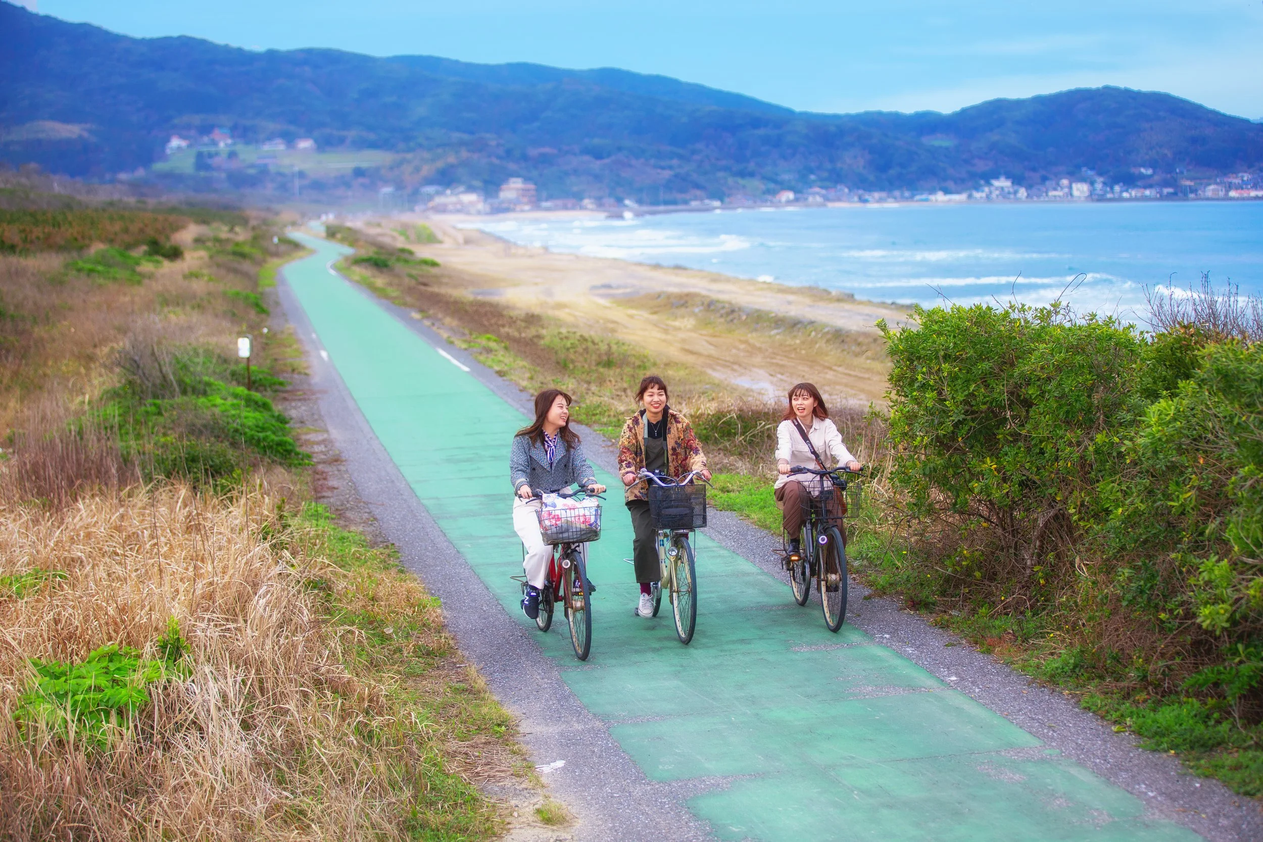 Three women cycling side-by-side on Okagaki's bright green coastal path beside white sandy beaches and distant hills in Fukuoka, Japan.