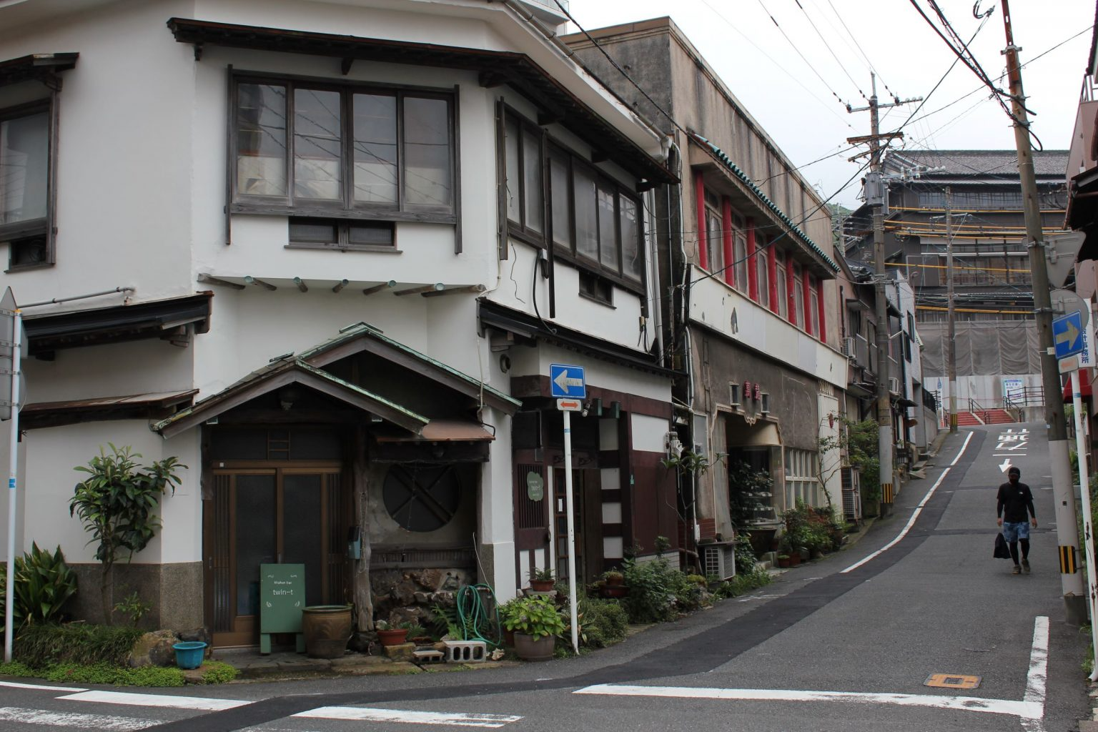 A narrow street on a hillside with old buildings on both sides, some with plants and outdoor items, and a person walking uphill with a bag.