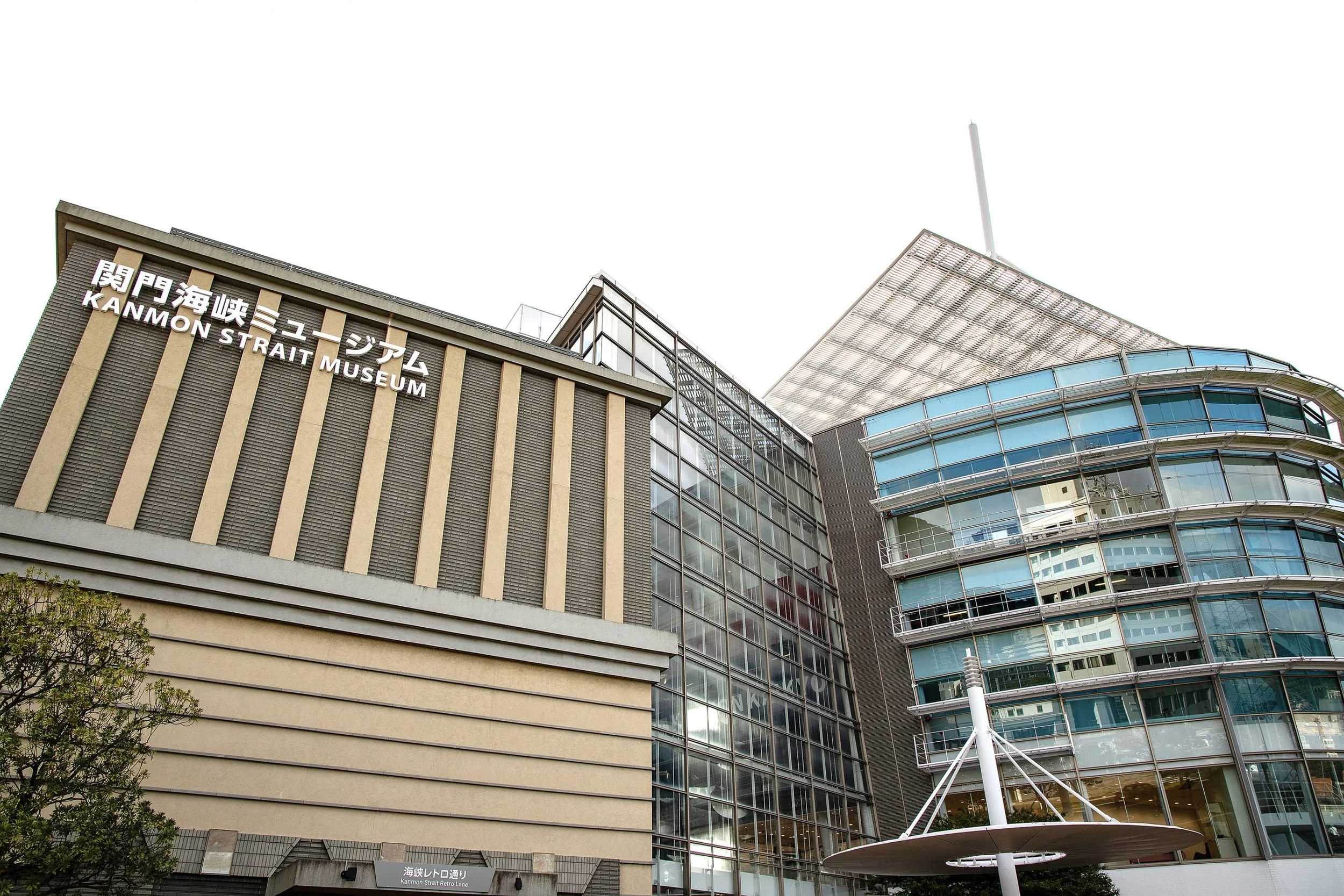 Exterior view of Kanmon Strait Museum building with modern architecture, glass windows, and a large umbrella structure nearby.