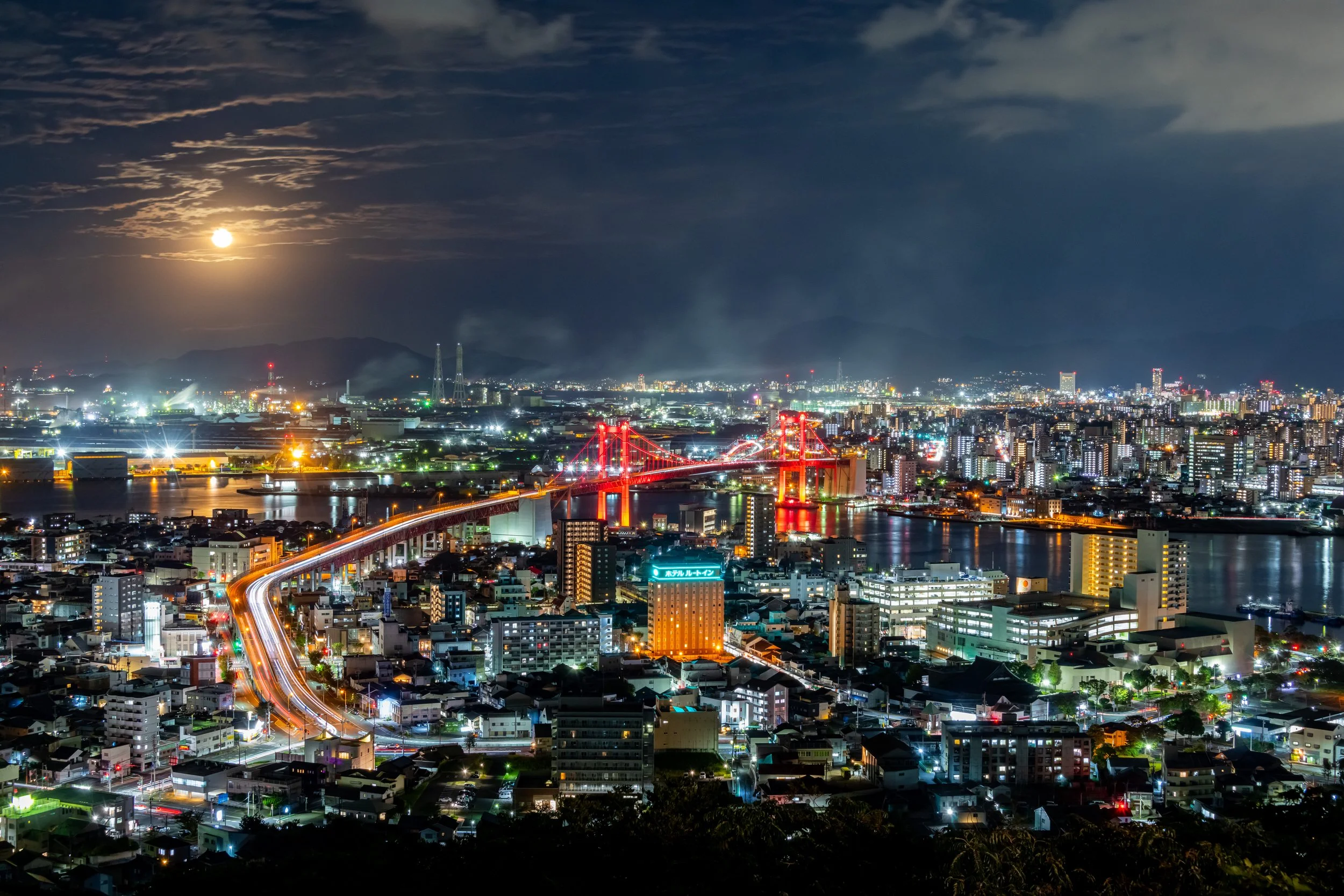 Panoramic night view from Takatoyama Park, Kitakyushu — the red-lit Wakato Bridge, glowing cityscape, and moonlit sky over Dokai Bay.