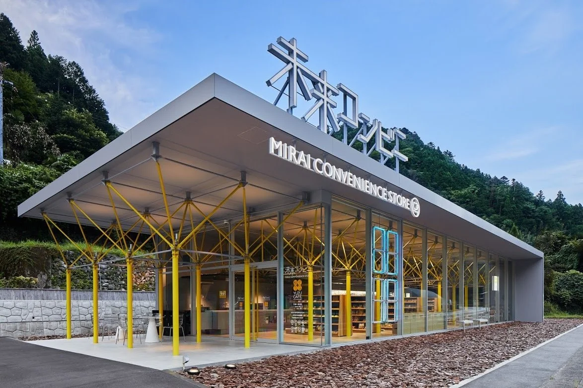 Glass-walled MIRAI Convenience Store framed by yellow steel columns against a forested hillside at dusk in Kito, Naka Town, Tokushima.