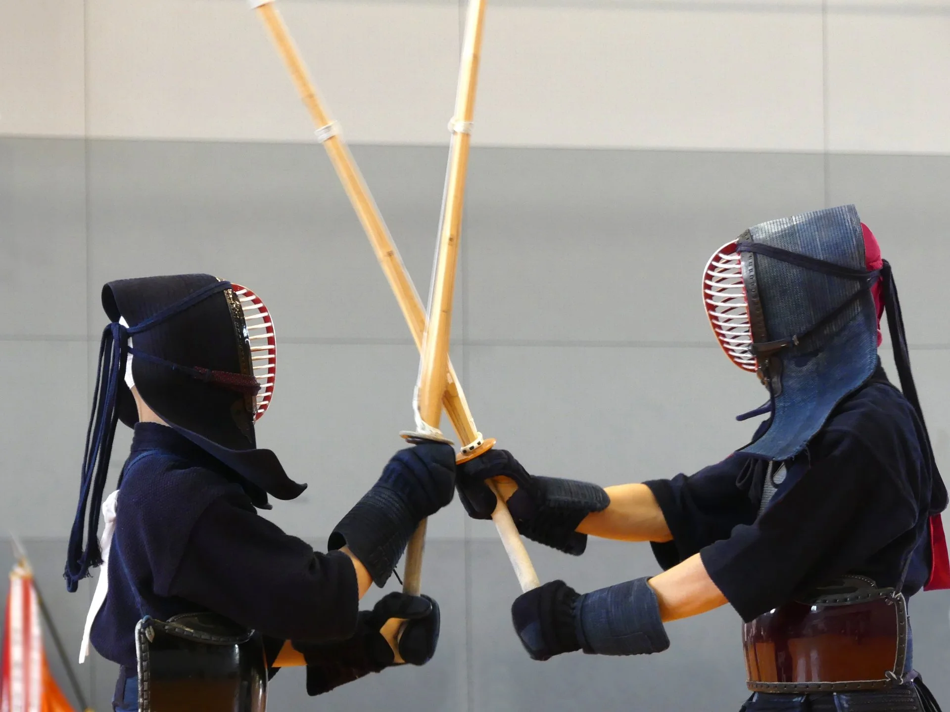 Two kendo practitioners wearing armor and helmets with face guards, practicing a match with wooden swords, inside a gym or dojo.