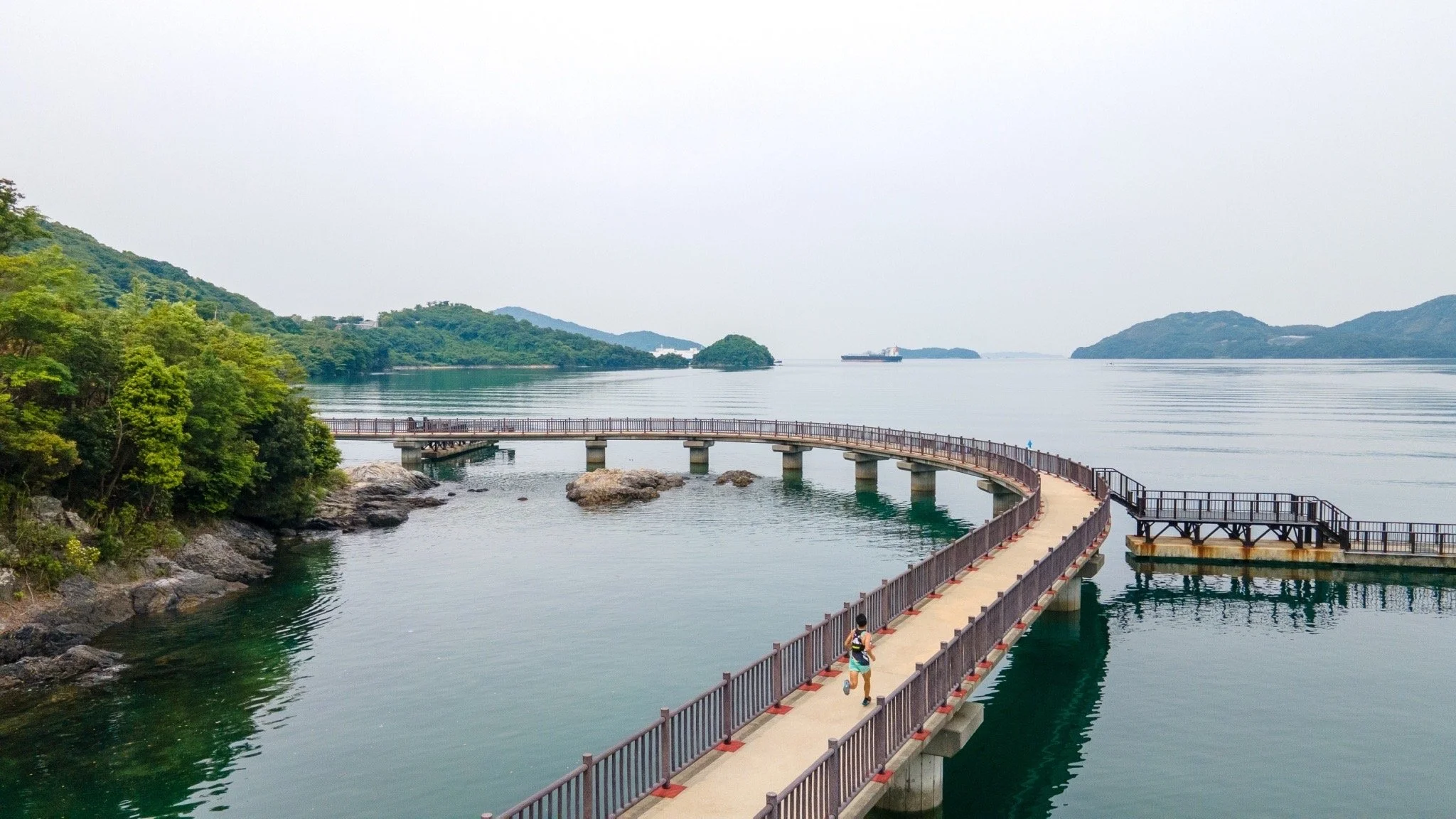 A lone jogger crosses a curved 300-meter sea walkway above the calm Seto Inland Sea in Kudamatsu, Yamaguchi — a hidden coastal gem.