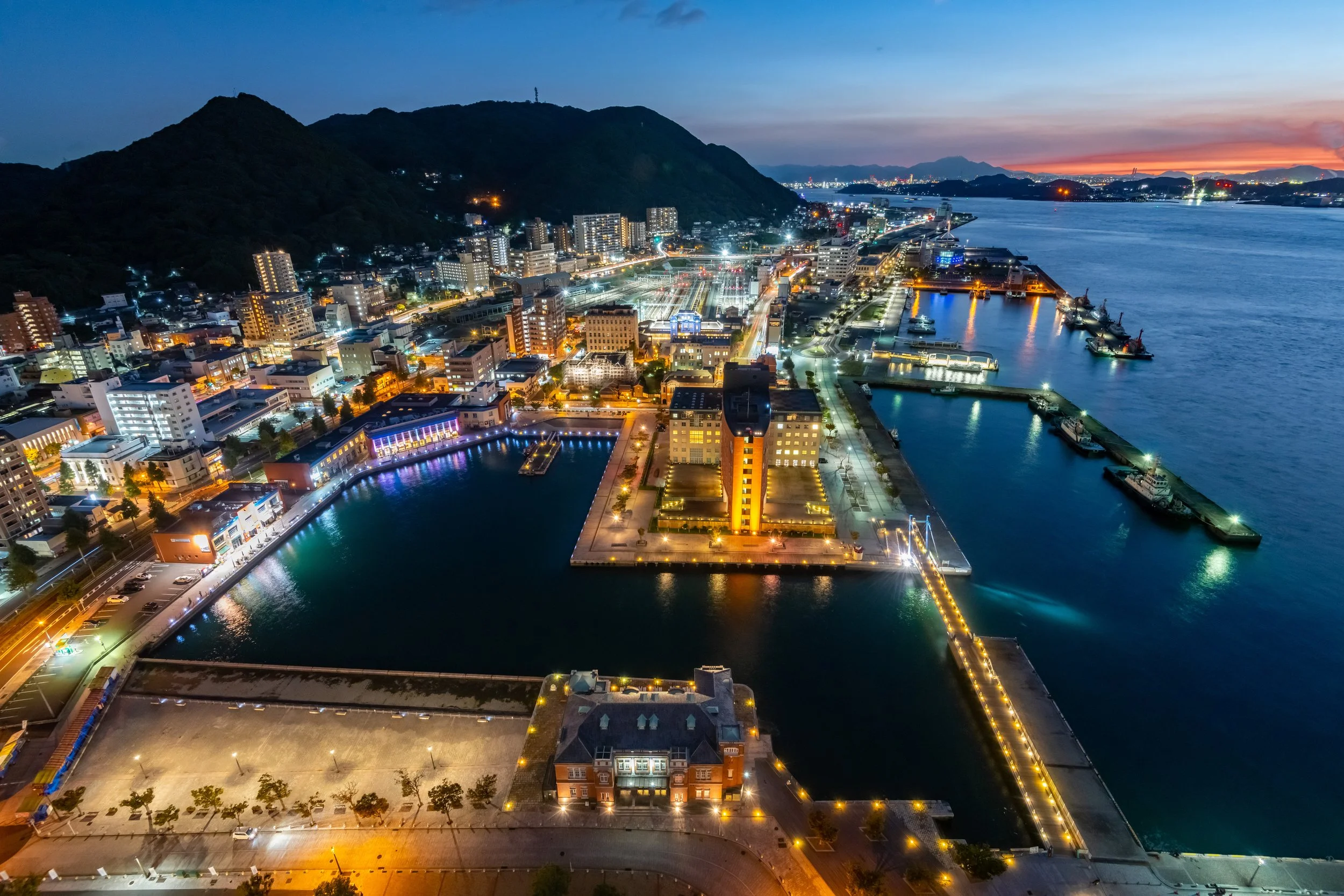 Aerial view of a cityscape at dusk with illuminated buildings, a harbor with boats, and a mountain in the background.