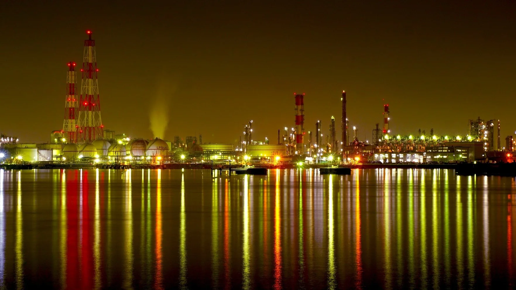 Takaishi factory nightscape glowing in red, green, and gold — LPG tanks and smokestacks reflected across the still bay waters of Osaka.