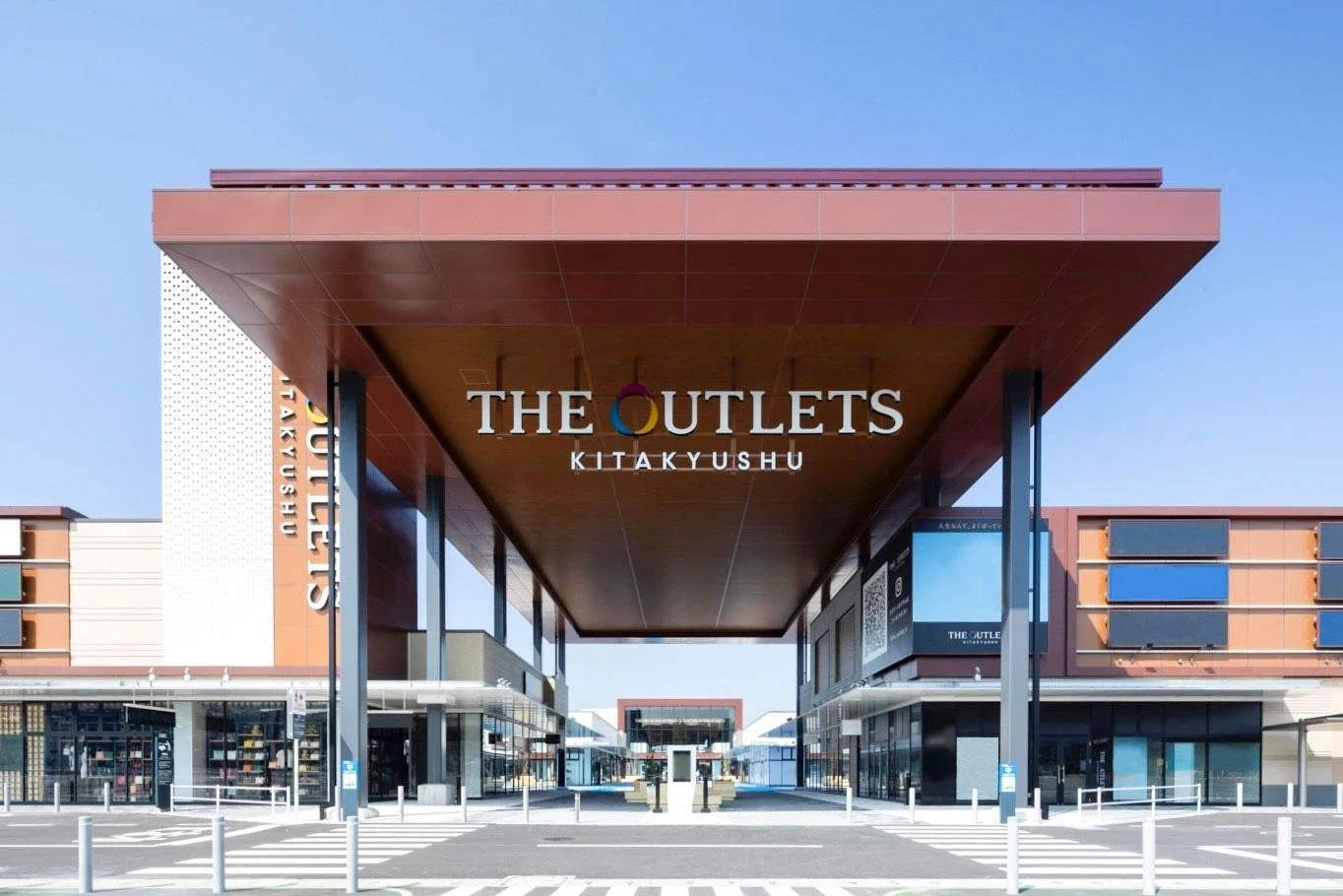 Front entrance of THE OUTLETS KITAKYUSHU, a large Kyushu shopping and entertainment complex with a modern terracotta canopy under clear blue sky.