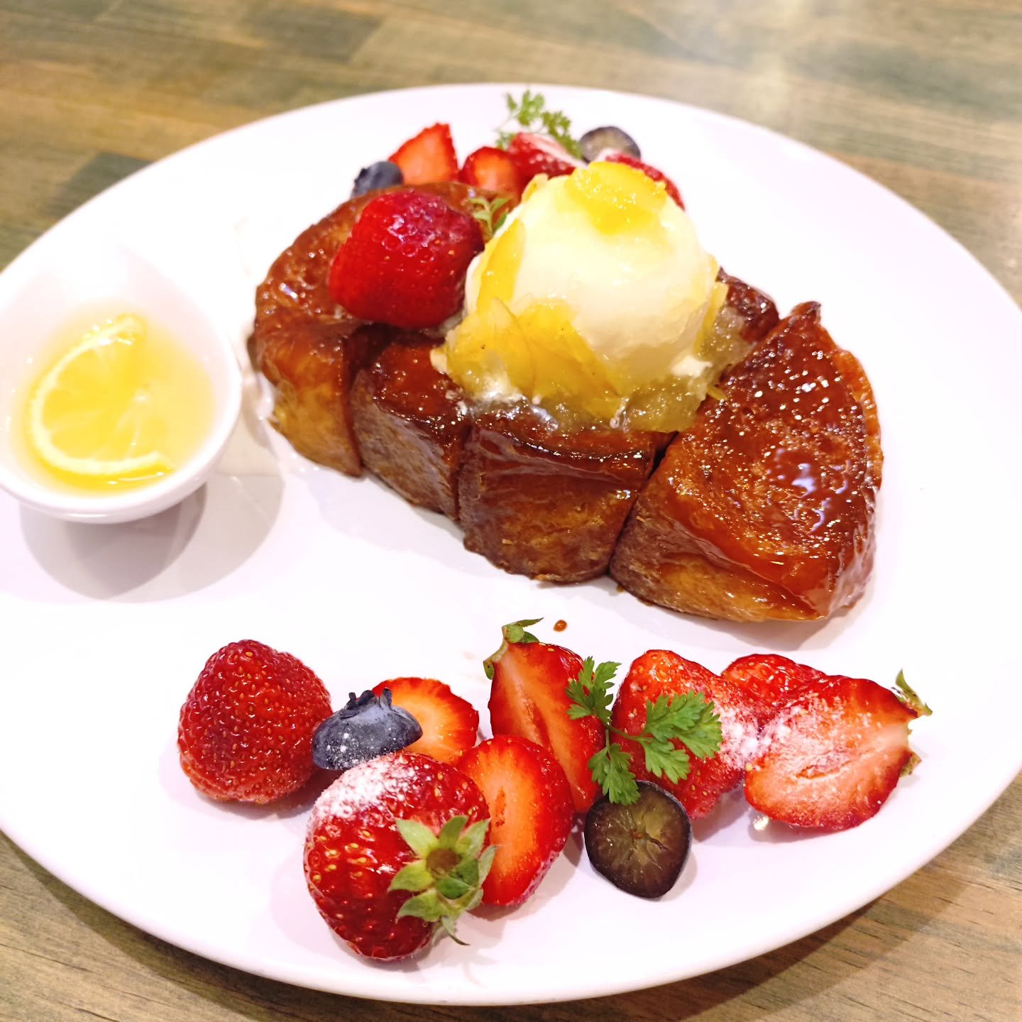 A plate of French toast topped with whipped cream, fresh strawberries, blueberries, and a scoop of vanilla ice cream with syrup. Garnished with mint leaves, with a small dish of lemon on the side.