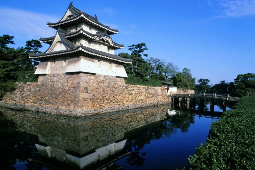 White-walled turret of Takamatsu Castle reflected in its seawater moat, with a wooden bridge leading across — a rare Japanese water castle in Tamamo Park, Kagawa.