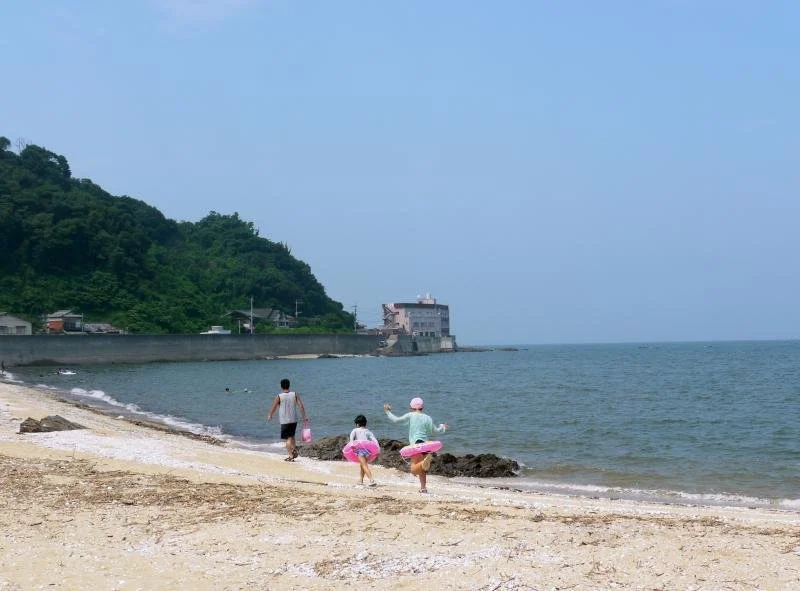 Families wading into the calm shallow waters of Minoshima Beach in Yukuhashi, Fukuoka, with a green coastal hill in the distance.