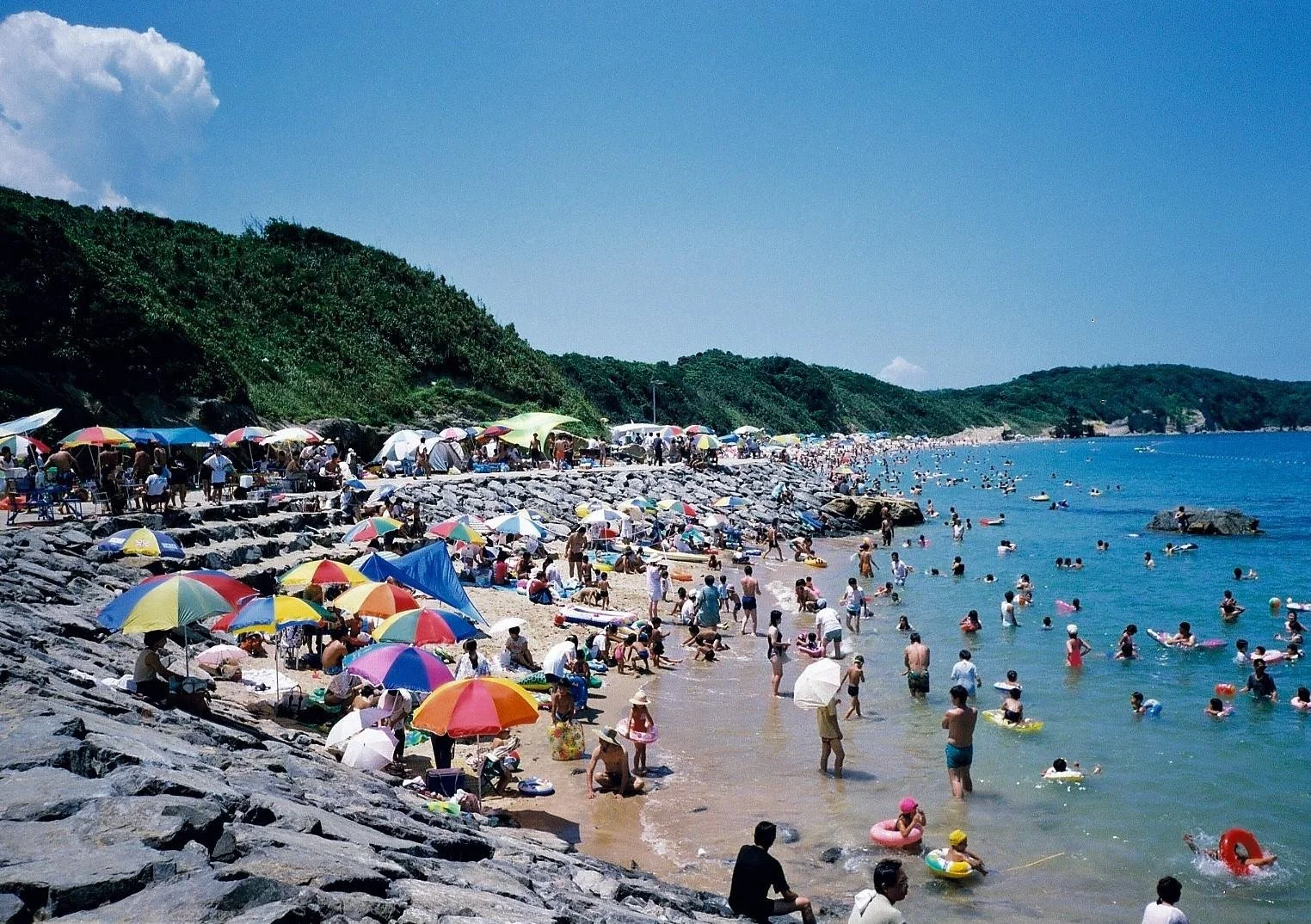 Colorful umbrellas line Waita Beach in Kitakyushu, families swimming in clear blue water framed by green headlands under bright summer sky.