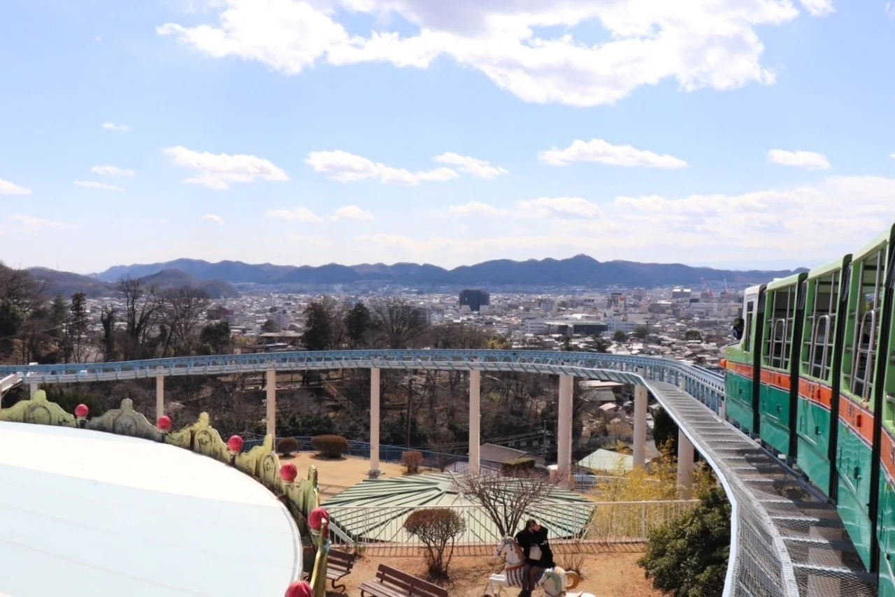 Elevated view from Kiryugaoka Amusement Park showing a retro green train on a curved track with Kiryu cityscape and distant mountains beyond.