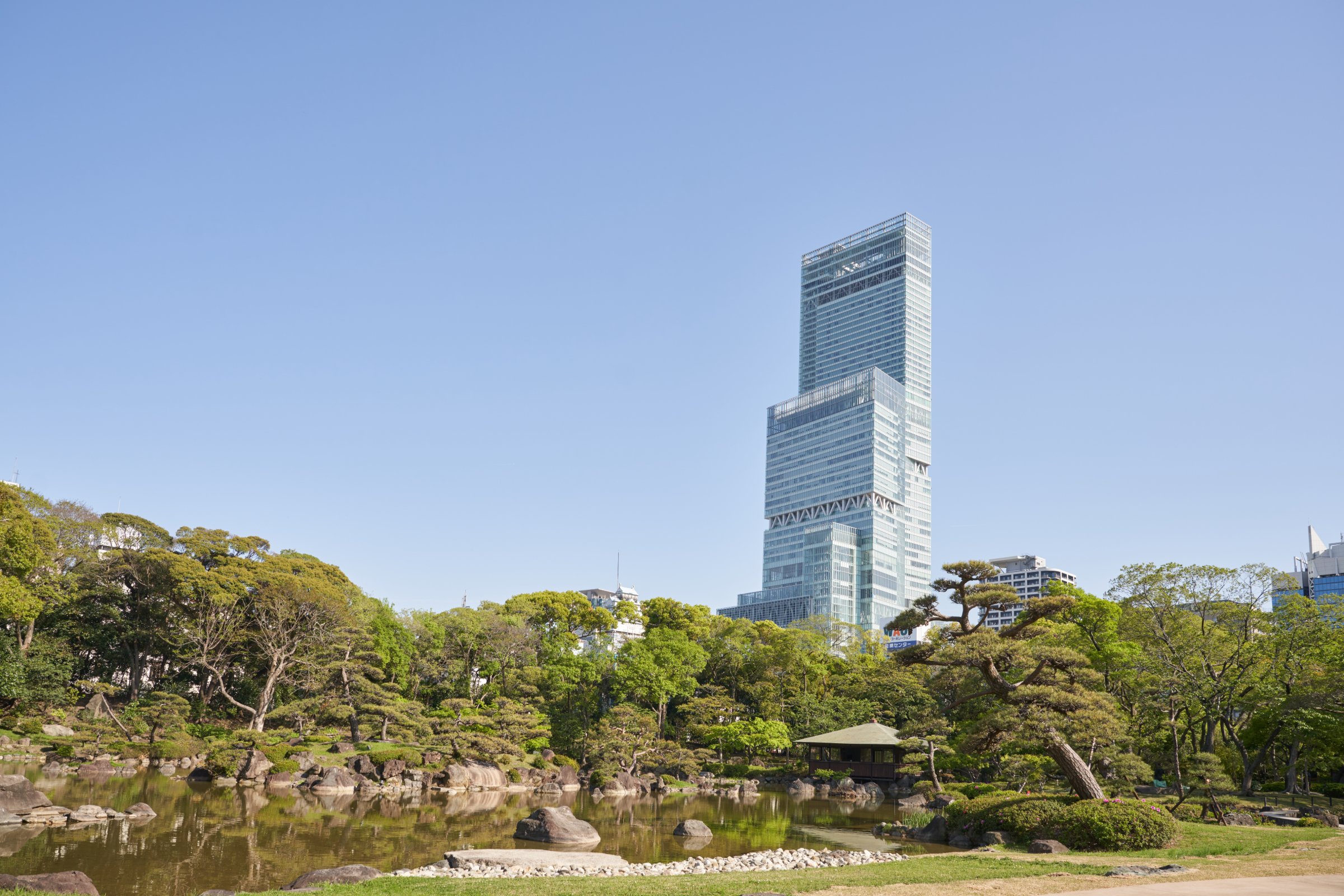 Traditional Japanese garden pond with sculpted pines and a wooden pavilion, Abeno Harukas skyscraper rising beyond the trees in Osaka.