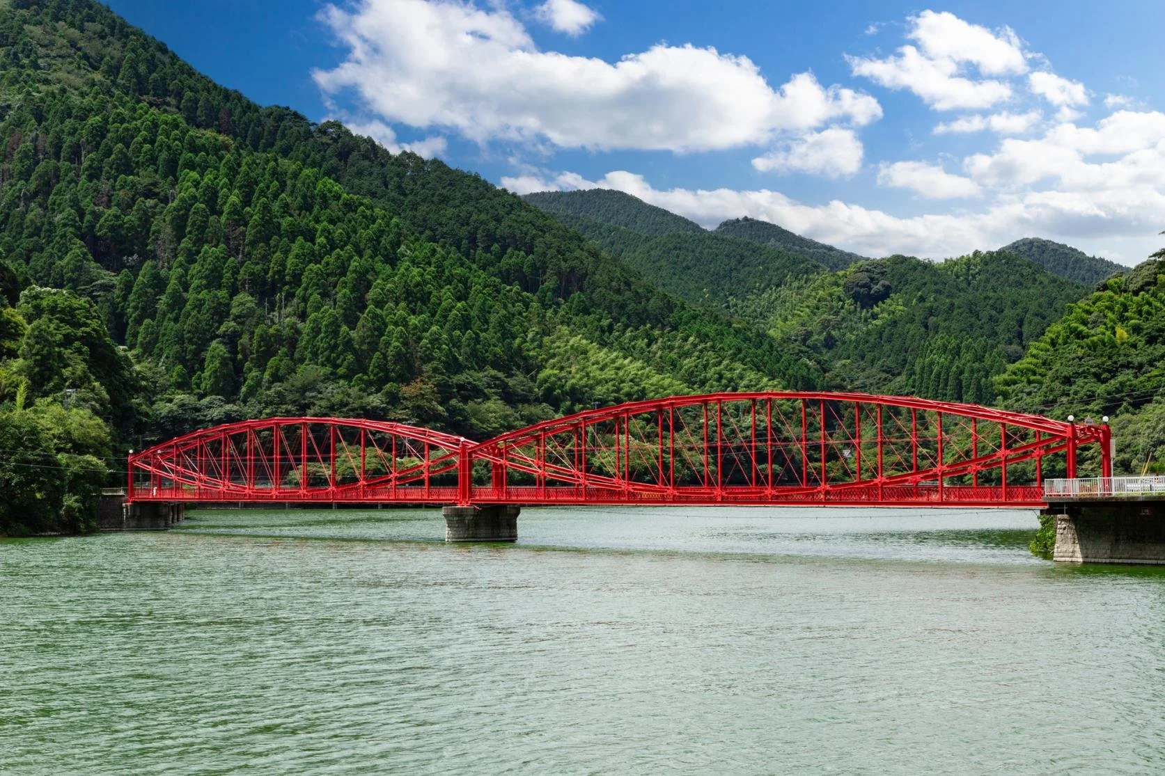 Red lenticular truss bridge arching over the emerald waters of Kawachi Reservoir, framed by forested hills in Kitakyushu, Japan.