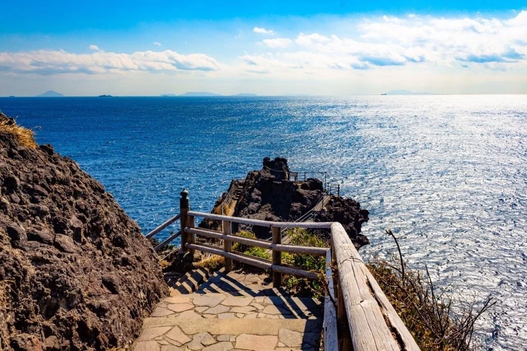 Stone trail with wooden railings stretching toward volcanic sea cliffs at Cape Irozaki — Japan's southernmost Pacific cape on a clear sunny day.