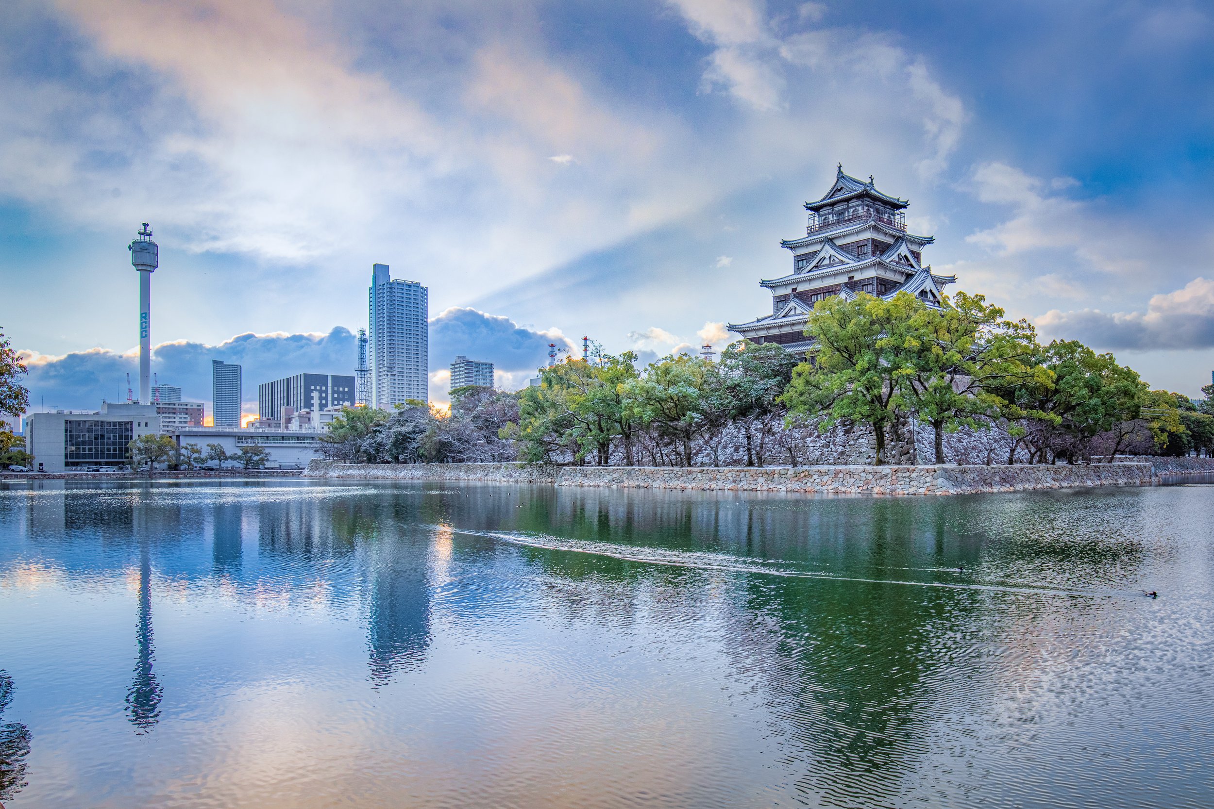 Hiroshima Castle's black keep mirrored on a still moat beneath a pastel dawn sky, framed by stone walls, trees, and the modern city skyline.
