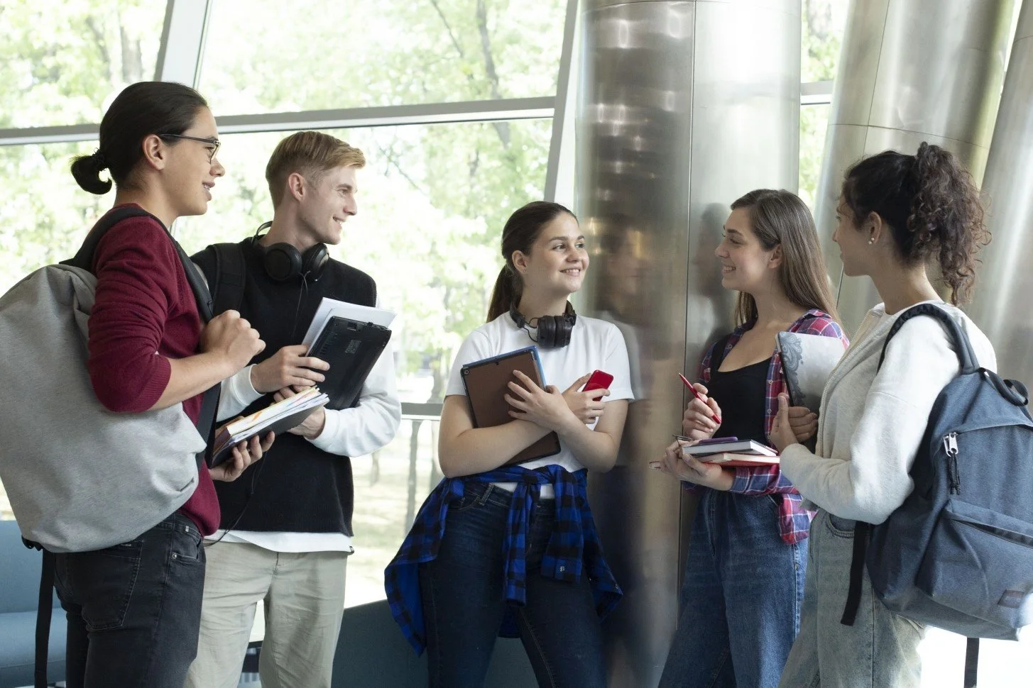 Five teenagers in an airport or transportation terminal, standing and chatting near a large window with trees visible outside. They are holding backpacks, notebooks, and electronic devices, and are engaged in conversation with smiling faces.