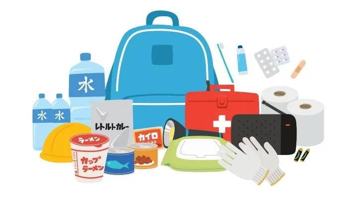Illustrated Japanese emergency preparedness kit: blue evacuation backpack, water bottles, canned food, first-aid kit, helmet, radio, and hygiene supplies.