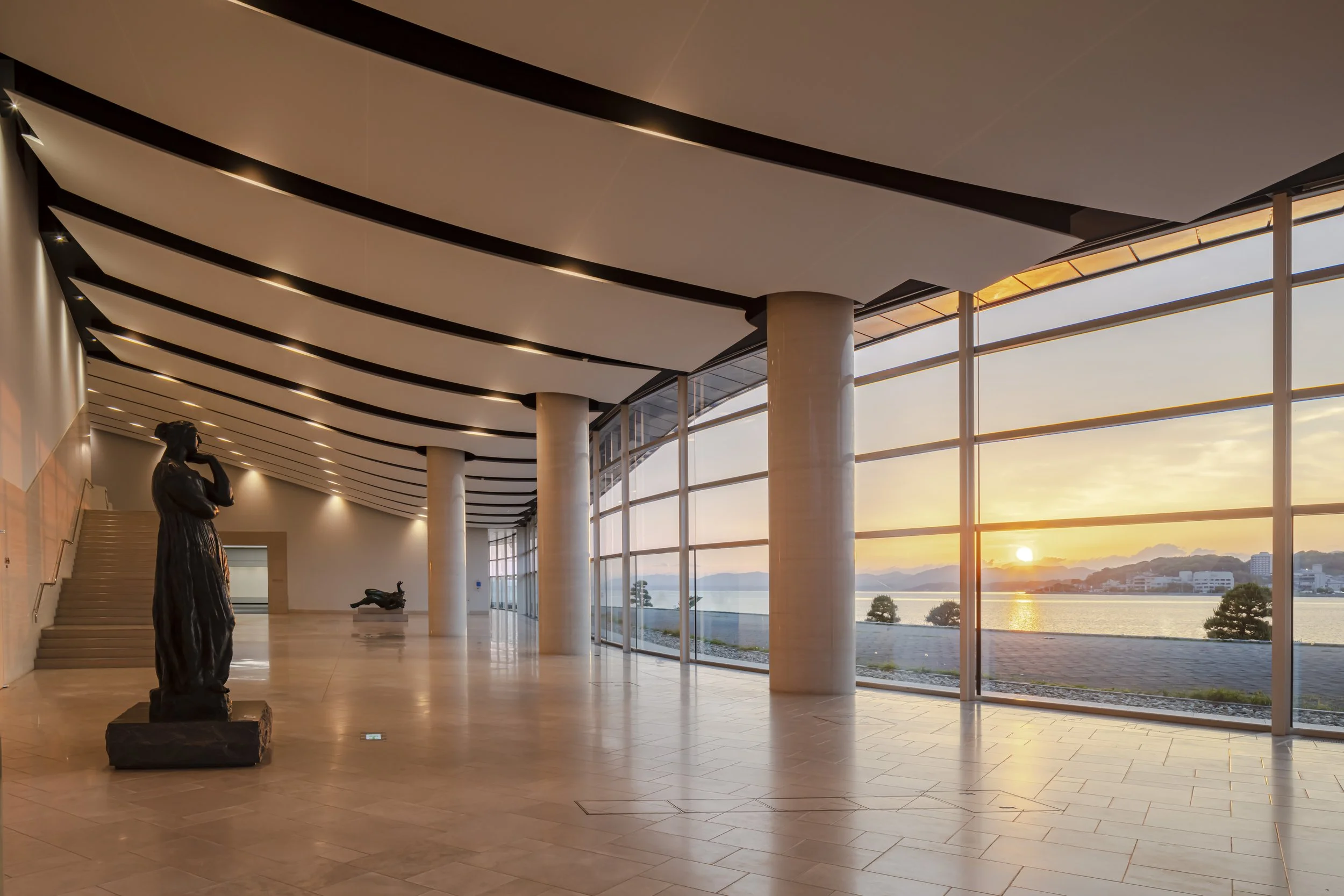 Bronze sculptures glowing in golden sunset light inside the glass lobby of Shimane Art Museum, overlooking Lake Shinji at dusk.