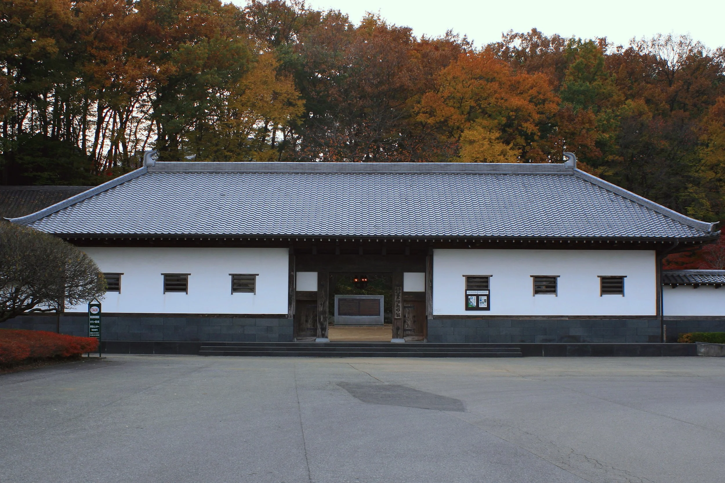 Traditional white-walled, tiled-roof entrance building at Kurita Museum in Ashikaga, framed by autumn foliage on the hillside behind.