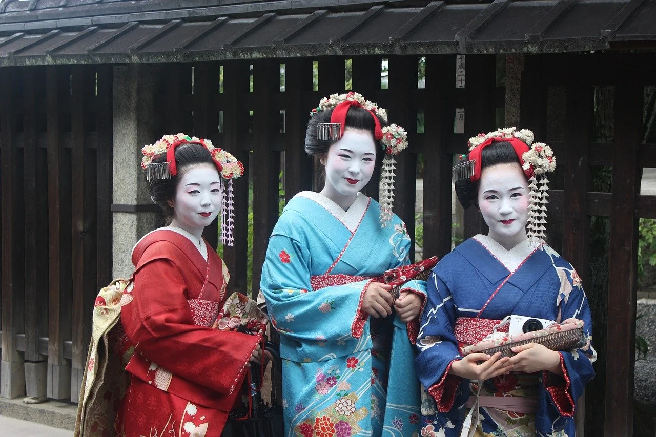 Three women dressed as geishas in traditional kimonos and white face makeup standing outdoors in front of a wooden fence.