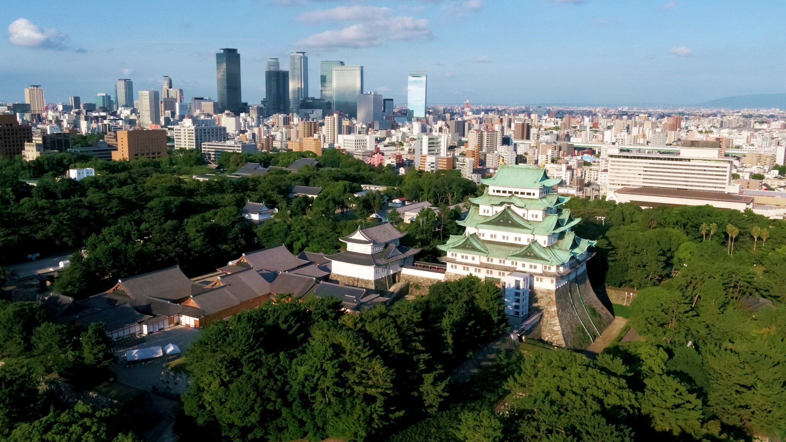 Aerial view of Nagoya Castle with green-tiled roofs and golden Kinshachi against the modern Nagoya skyline under a blue sky.
