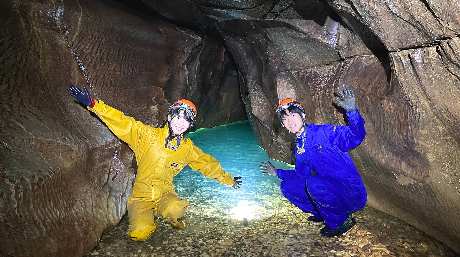 Two people wearing yellow and blue waterproof suits and helmets with headlamps, kneeling inside a narrow cave or tunnel with smooth brown rock walls and a small body of water.