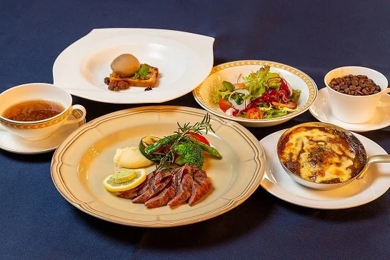 Assorted dishes including salad, grilled meat with lemon and broccoli, baked dish, cup of coffee, and a bowl of black beans on a dark table.