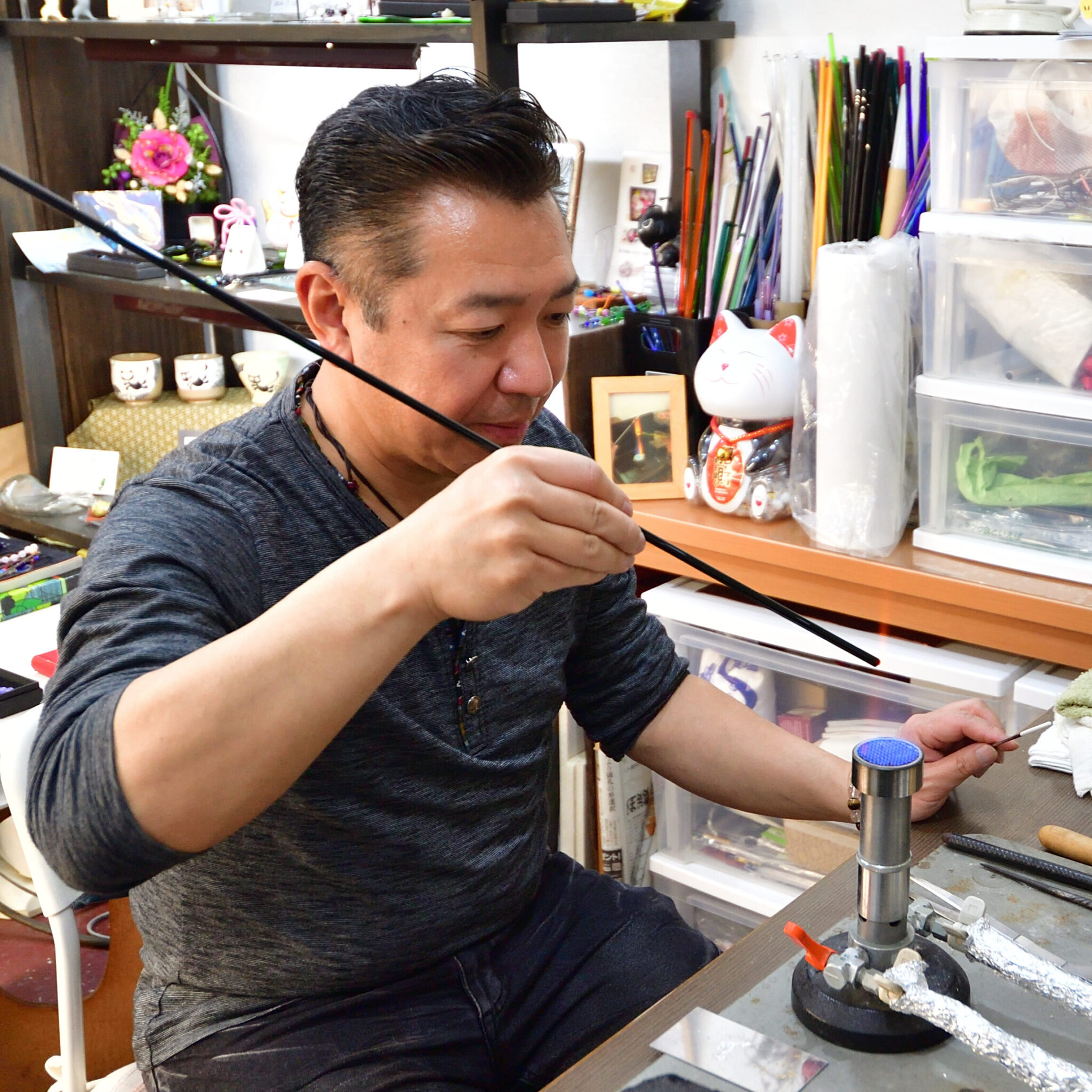 A man working carefully on an art project at a cluttered workspace, with various art supplies and decorative items in the background.