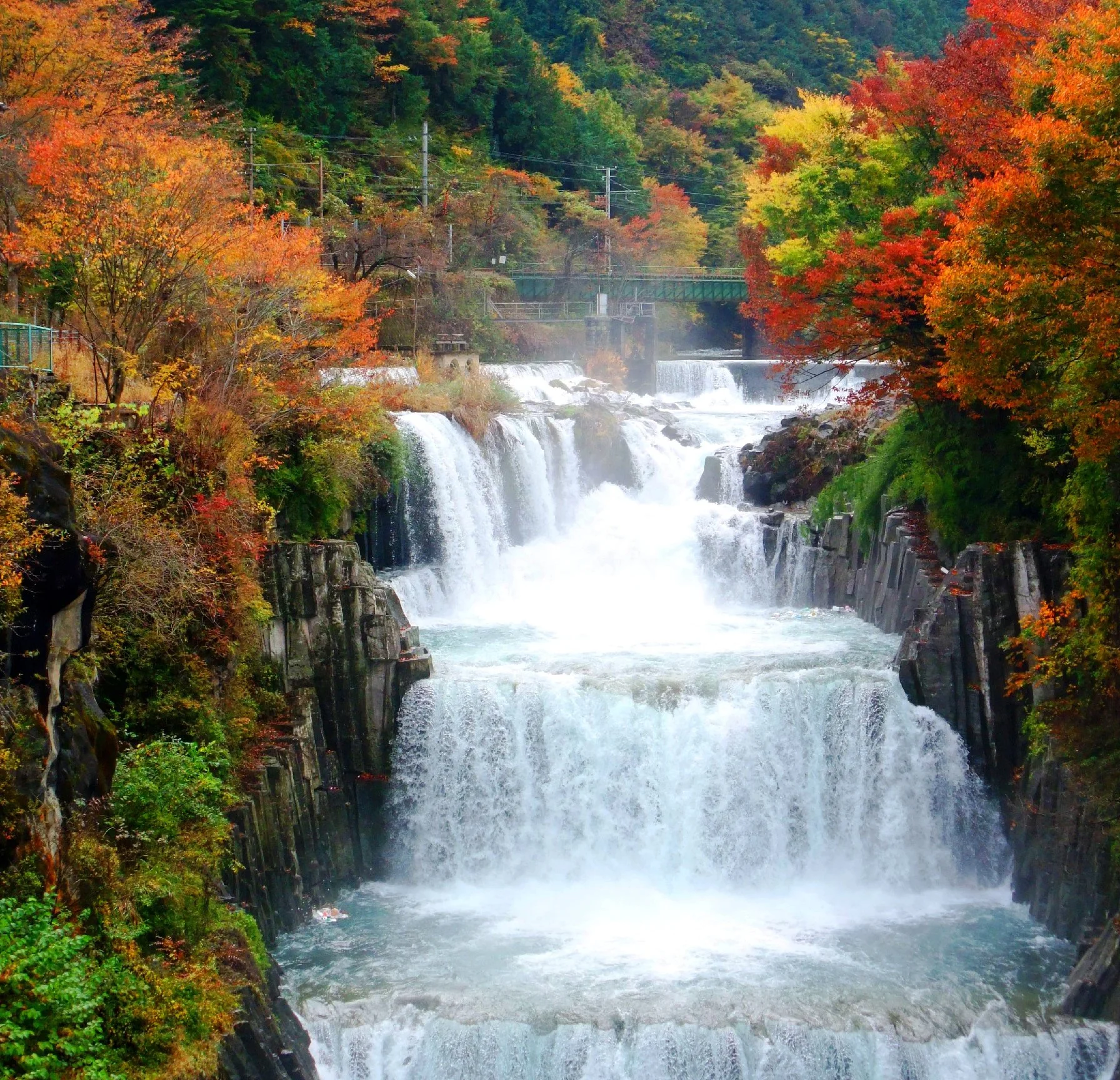 Meisho Tahara Falls cascading through a rocky gorge framed by vivid autumn foliage in Tsuru, Yamanashi — a hidden sanctuary near Mount Fuji.